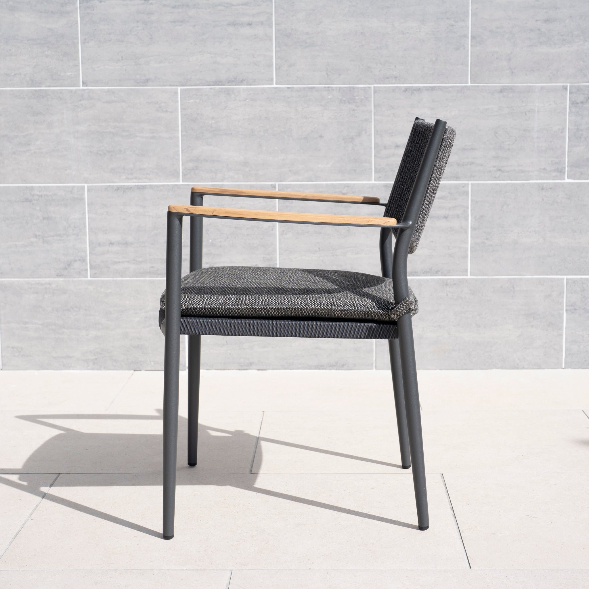 The Tellaro Rope Dining Chair in Grey (6pcs) features wooden armrests and a weather-resistant cushion, displayed on a light stone patio with a light gray tiled wall in the background.