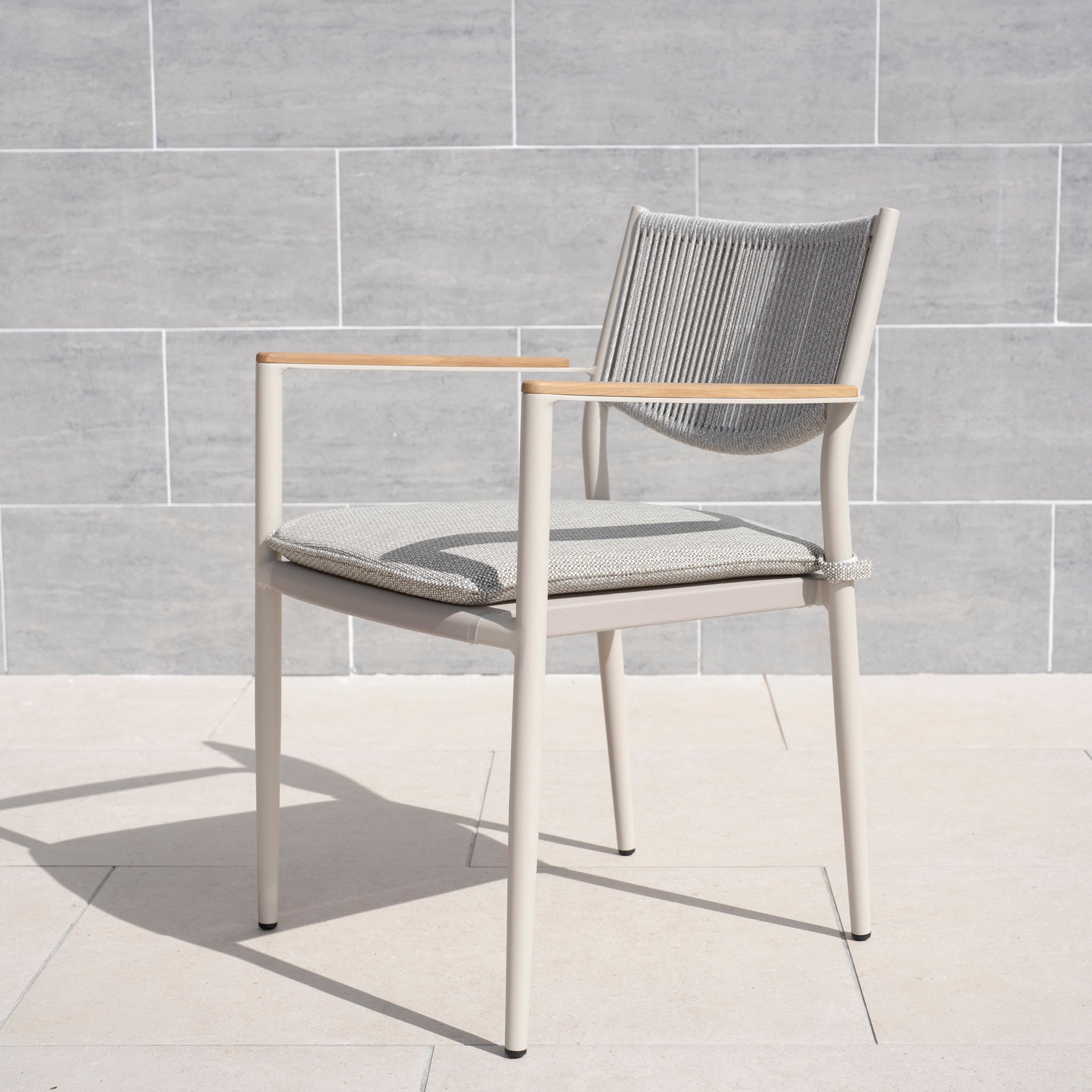 A Tellaro Rope Dining Chair in Latte (set of 6), featuring a hand-woven rope back and light wood armrests, sits on a tiled patio by a gray stone wall.