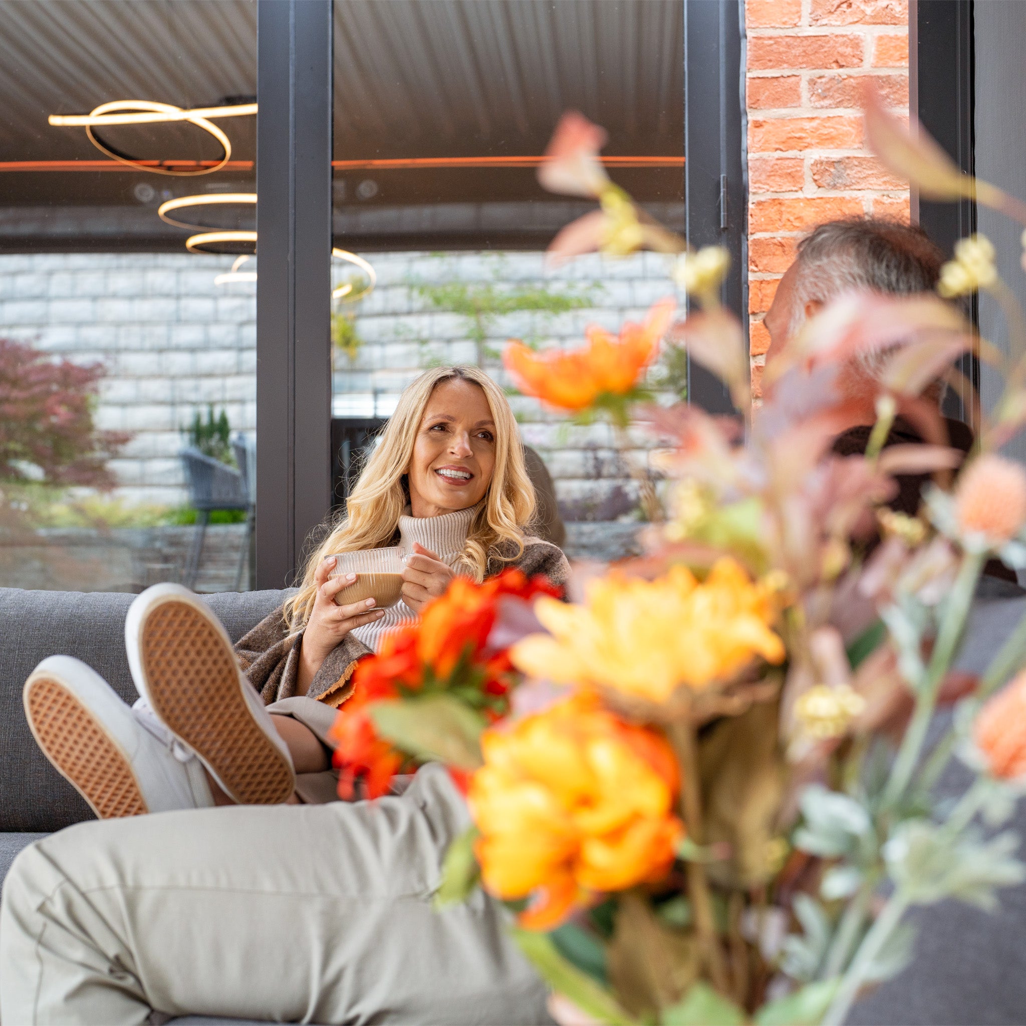 A woman sits on a sofa with a mug, smiling. In the foreground, blurred flowers are near large windows fitted with PergoSTET Plus 3m x 4m Rectangular Pergola in Grey, featuring 3 drop sides and LED lighting.
