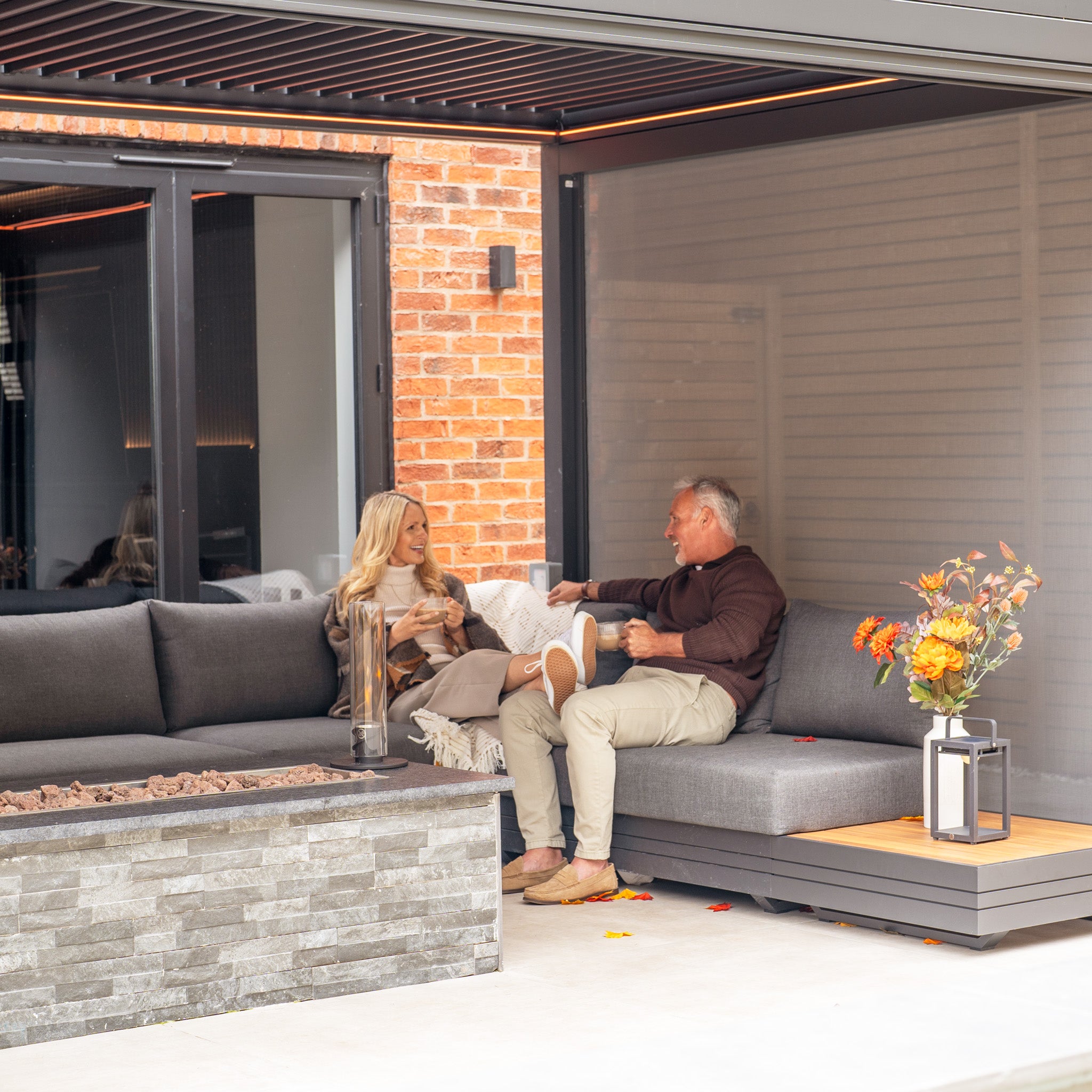 A couple sits on an outdoor couch under a sleek PergoSTET Plus 3m x 4m pergola with 3 drop sides and LED lighting in grey, their conversation dancing in the firelight. They hold mugs, enveloped in the cozy ambiance of their modern patio setting.