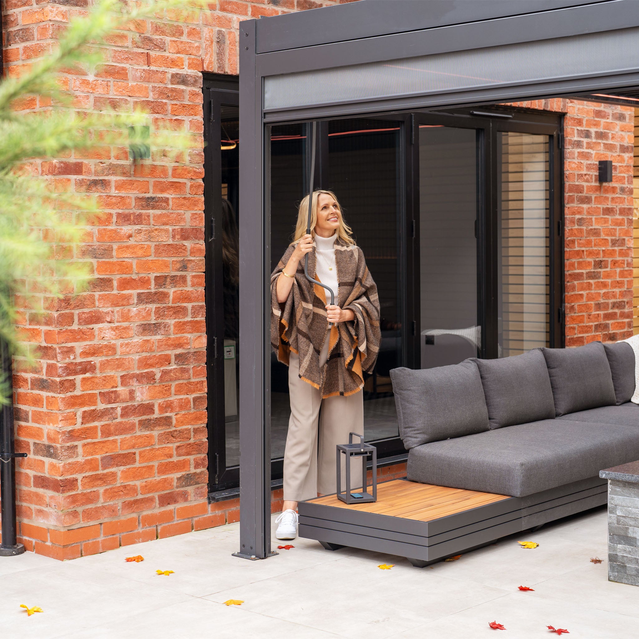 A woman in a cozy shawl stands on a patio beneath the PergoSTET Plus 3m x 4m Rectangular Pergola with 3 Drop Sides and LED Lighting in Grey, next to an outdoor sofa and lantern, surrounded by brick walls and fallen leaves.