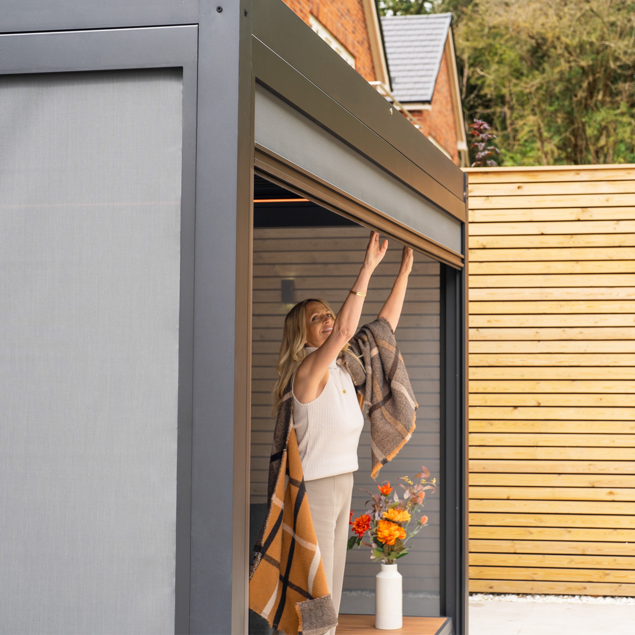 Wrapped in a plaid blanket, the person skillfully adjusts the awning of the PergoSTET Plus 3m x 4m Rectangular Pergola with 3 Drop Sides and LED Lighting in Grey, transforming it into an inviting space surrounded by vibrant flowers in a vase.