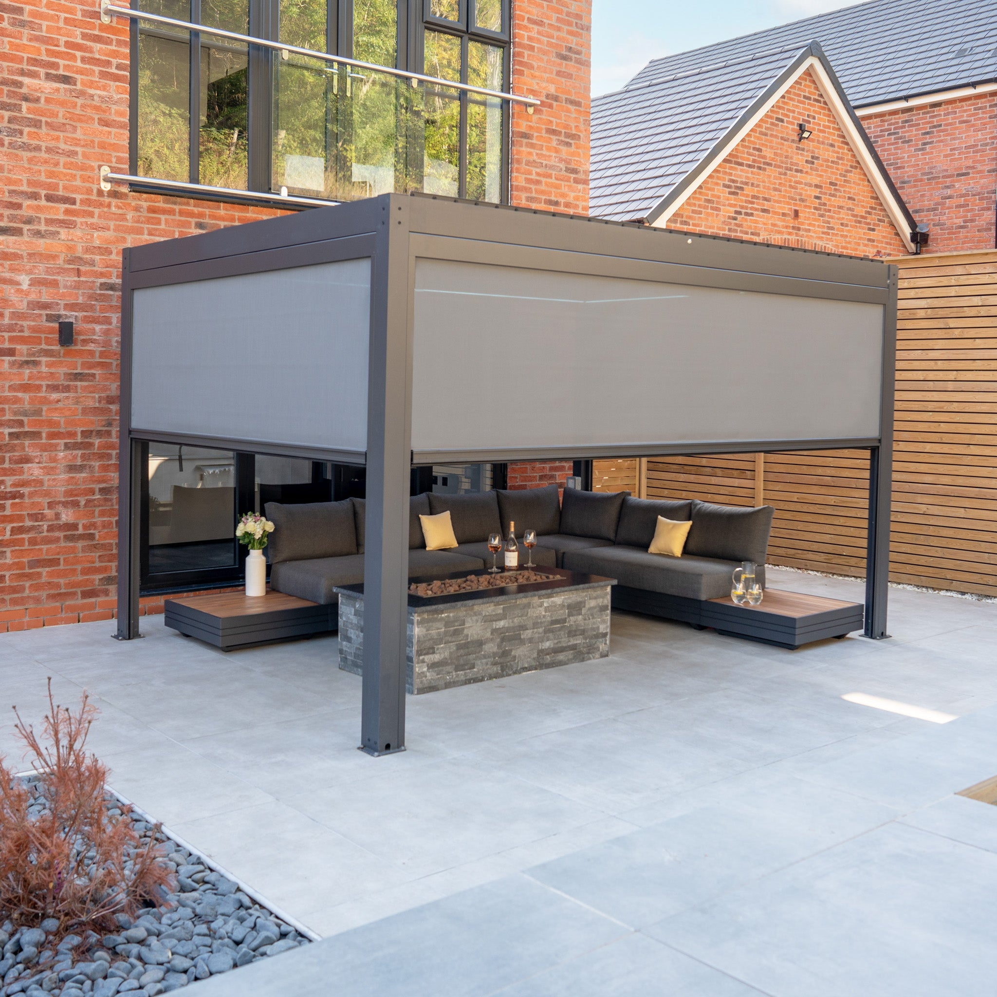 Modern outdoor patio featuring the sleek PergoSTET Plus 3m x 4m Rectangular Pergola in grey, complete with three manual side blinds and integrated LED lighting. The space is complemented by a stylish sectional sofa, coffee table, and decorative pillows—perfect for relaxing beneath a modern pergola designed for both comfort and style.