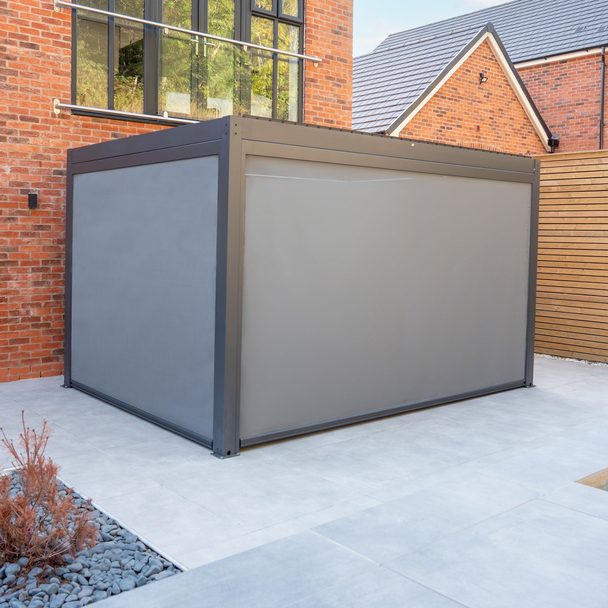 A PergoSTET Plus 3m x 4m rectangular pergola in grey, equipped with three manual side blinds and integrated LED lighting, stands on a tiled patio beside a brick house, offering modern comfort and style.