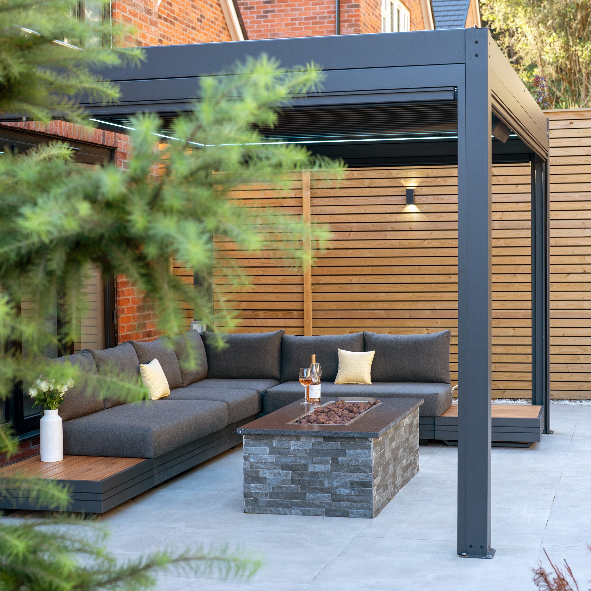 A modern patio features a gray sectional sofa, a stone fire pit, and a wooden fence partly obscured by tree branches. The space is accentuated by the PergoSTET Plus 3m x 4m Rectangular Pergola with LED Lighting in Grey, offering a blend of shade and style.