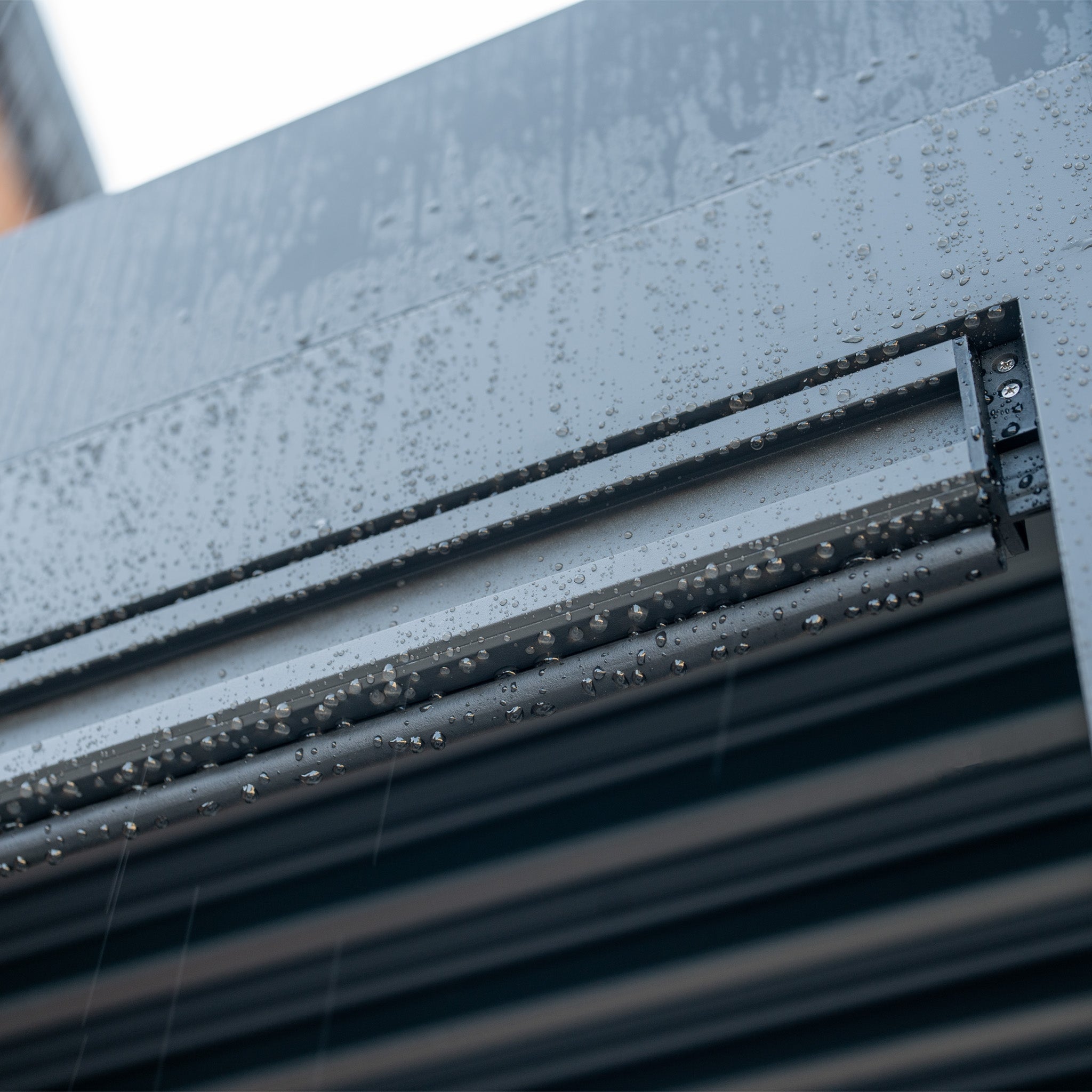 Raindrops bead on the sleek surface of a PergoSTET Plus 3m x 4m Rectangular Pergola with 3 Drop Sides and LED Lighting in Grey, reminiscent of nature's dance on metallic structures.