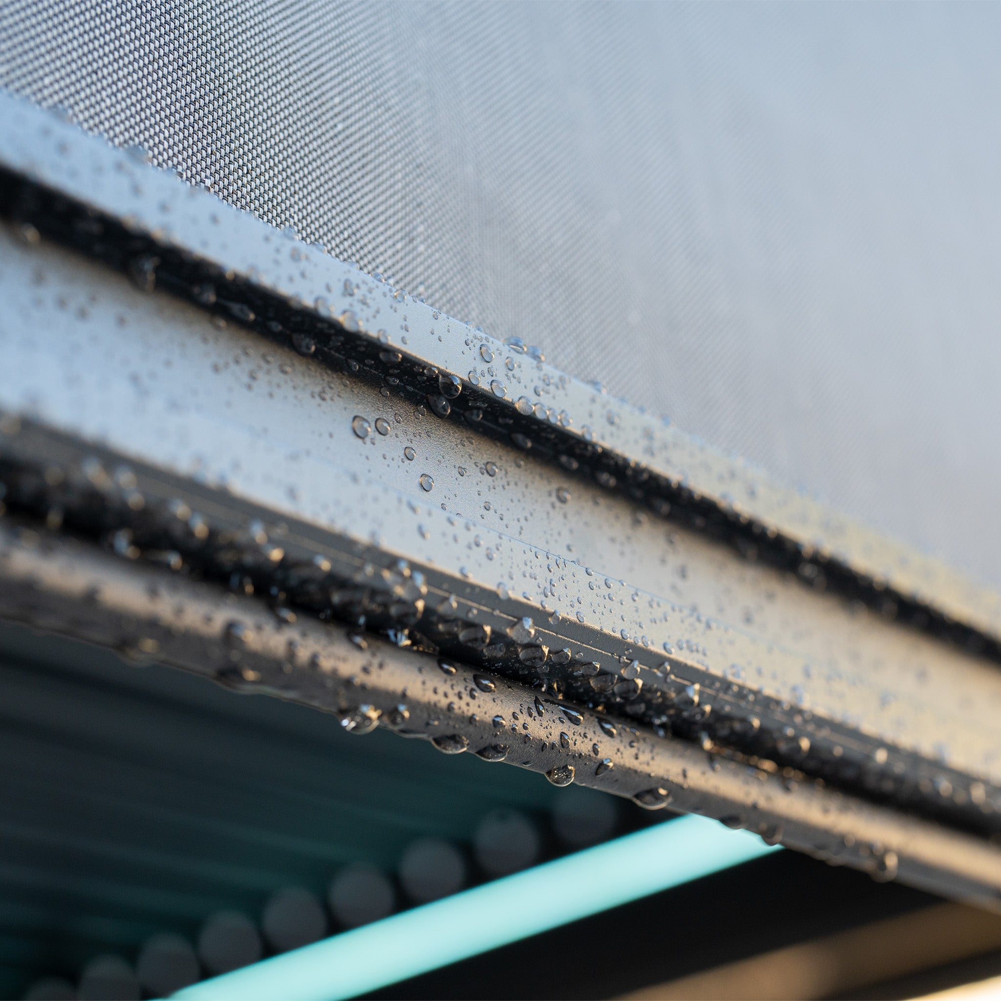 Close-up of raindrops sliding down a sleek metal surface, set against the perforated backdrop of a PergoSTET Plus 3m x 4m Rectangular Pergola with 3 Drop Sides and LED Lighting in Grey.