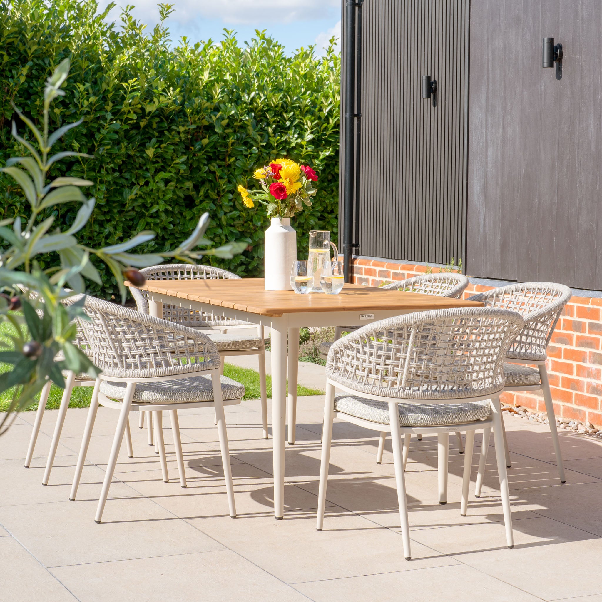 Presenting the Kalama 6 Seat Rectangular Dining Set with Teak Table in Latte: an outdoor patio collection that includes a sophisticated teak table and six woven chairs. A vase with fresh flowers beautifully complements the set, surrounded by a brick wall and vibrant greenery. Crafted with weather-proof aluminum, this ensemble is stylish and robust for all seasons.