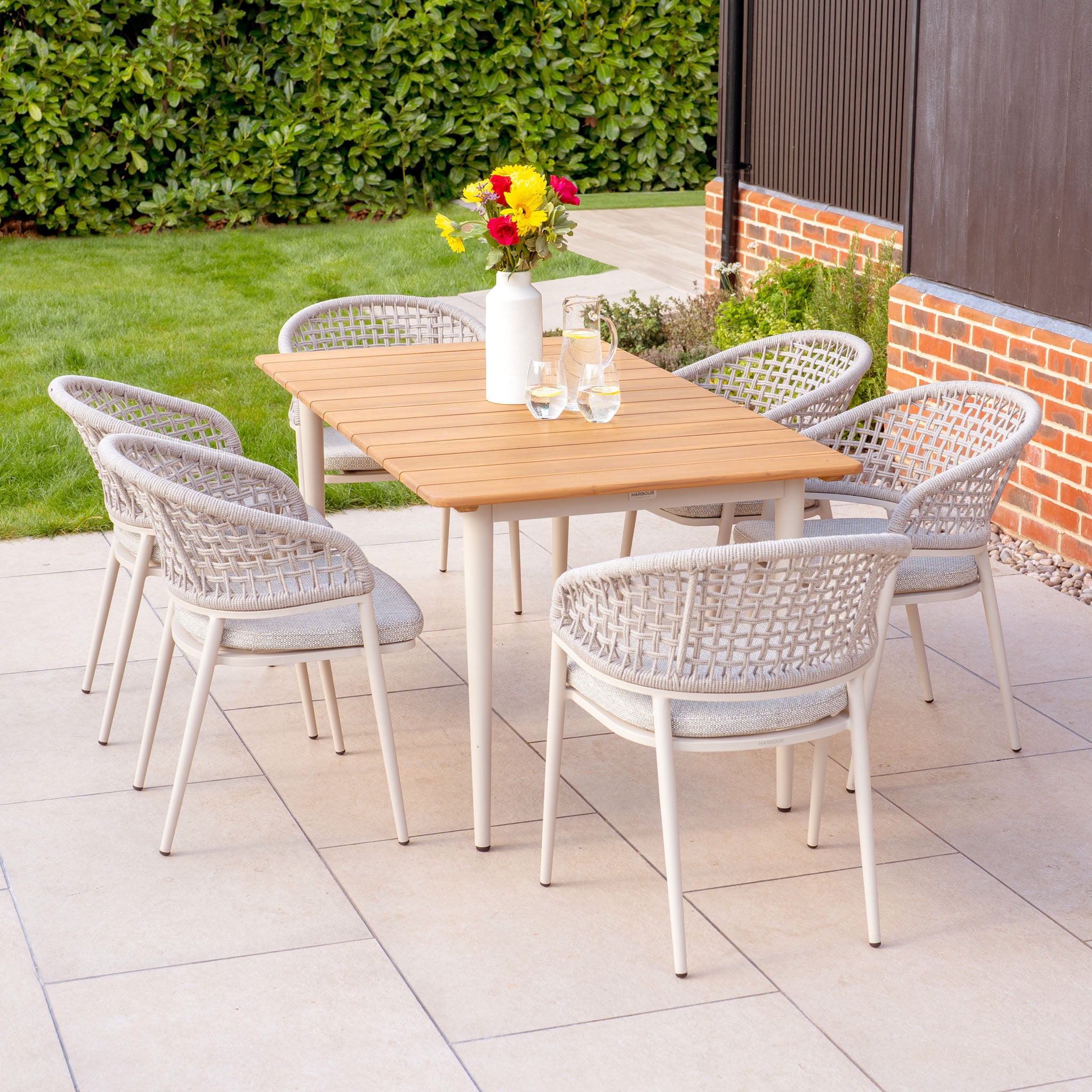 The Kalama 6 Seat Rectangular Dining Set with Teak Table in Latte includes six wicker chairs and a teak table, ideal for outdoor dining. Decorated with flowers and glasses, this weather-proof aluminum set enhances any patio adjacent to a lawn and brick wall.