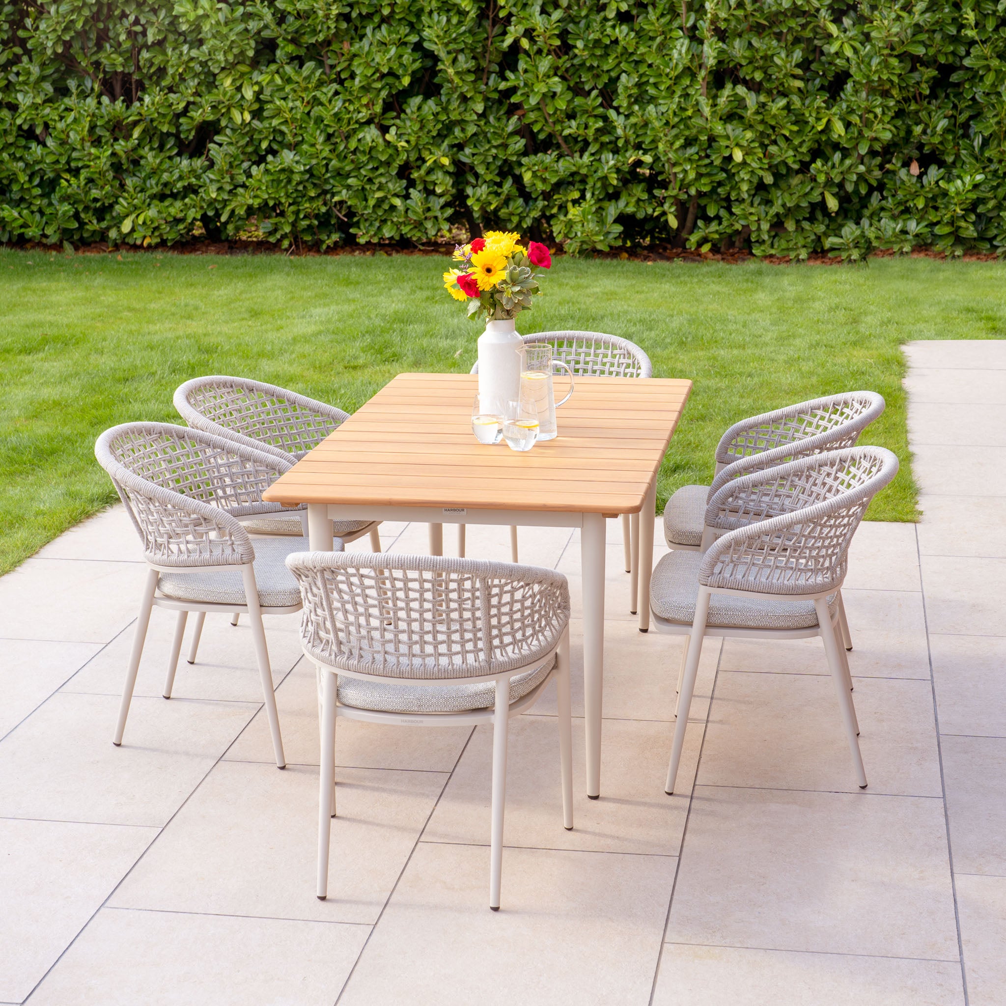 The Kalama 6 Seat Rectangular Dining Set with Teak Table in Latte boasts a sleek rectangular teak table accompanied by six wicker chairs, making it ideal for your patio. This elegant arrangement is accented with a white vase of flowers and constructed from weatherproof aluminum, ensuring long-lasting outdoor enjoyment.