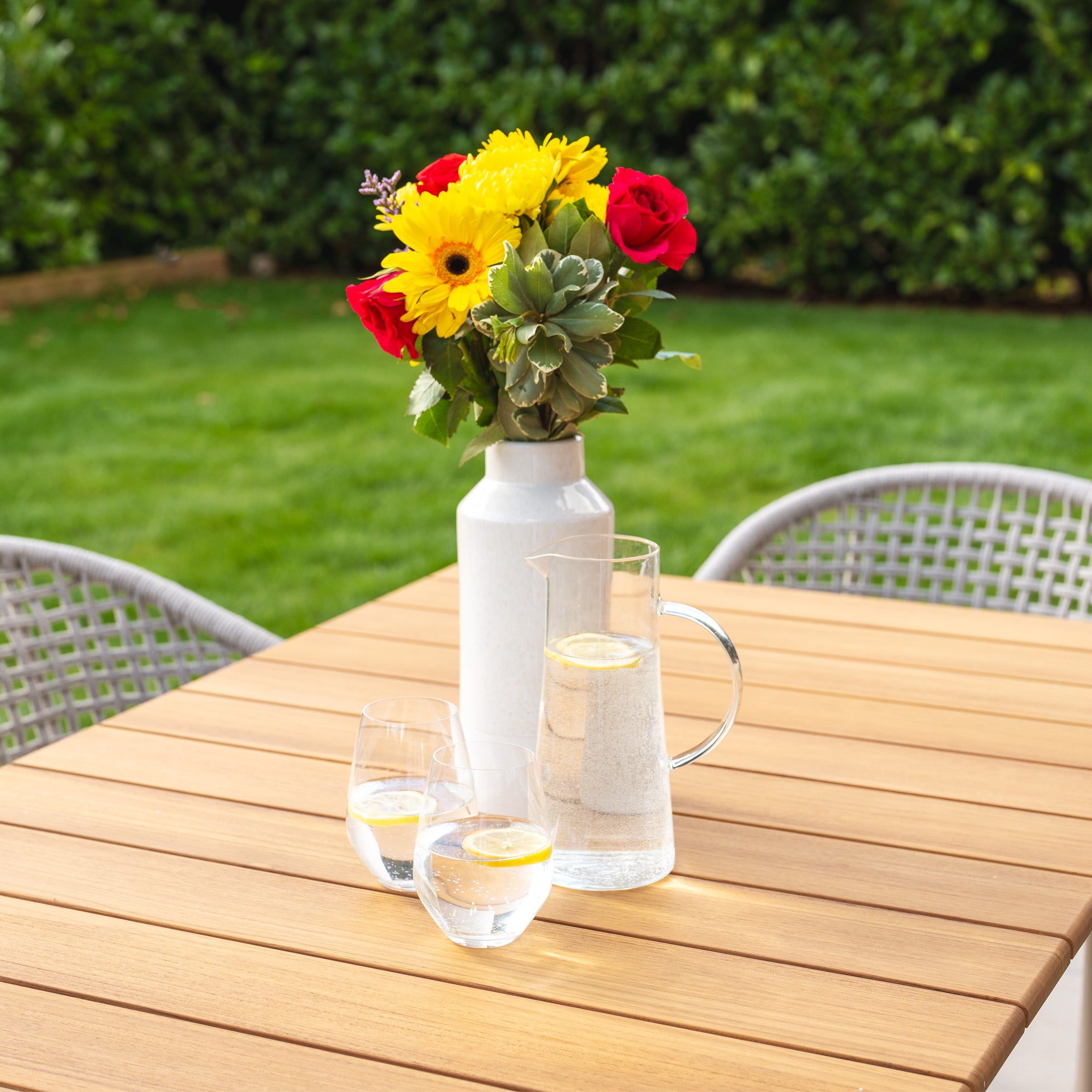 A bouquet in a white vase, a pitcher of lemon water, and two glasses rest on the Kalama 6 Seat Rectangular Dining Set's teak table in Latte, accompanied by chairs and greenery to enhance the wooden patio's charm.