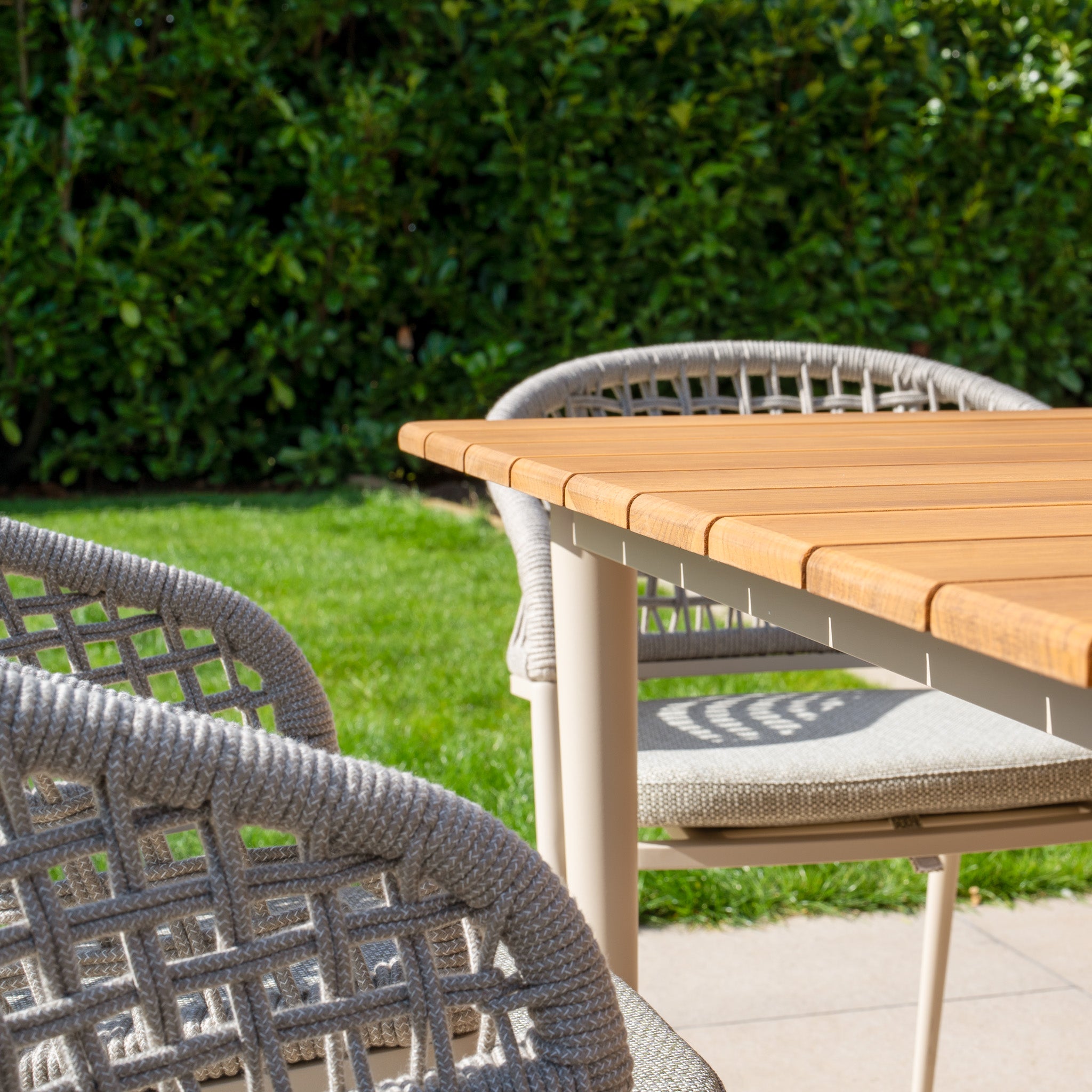 The Kalama 6 Seat Rectangular Dining Set with Teak Table in Latte, featuring a weatherproof aluminum frame, sits elegantly on a tiled surface, enveloped by lush greenery.