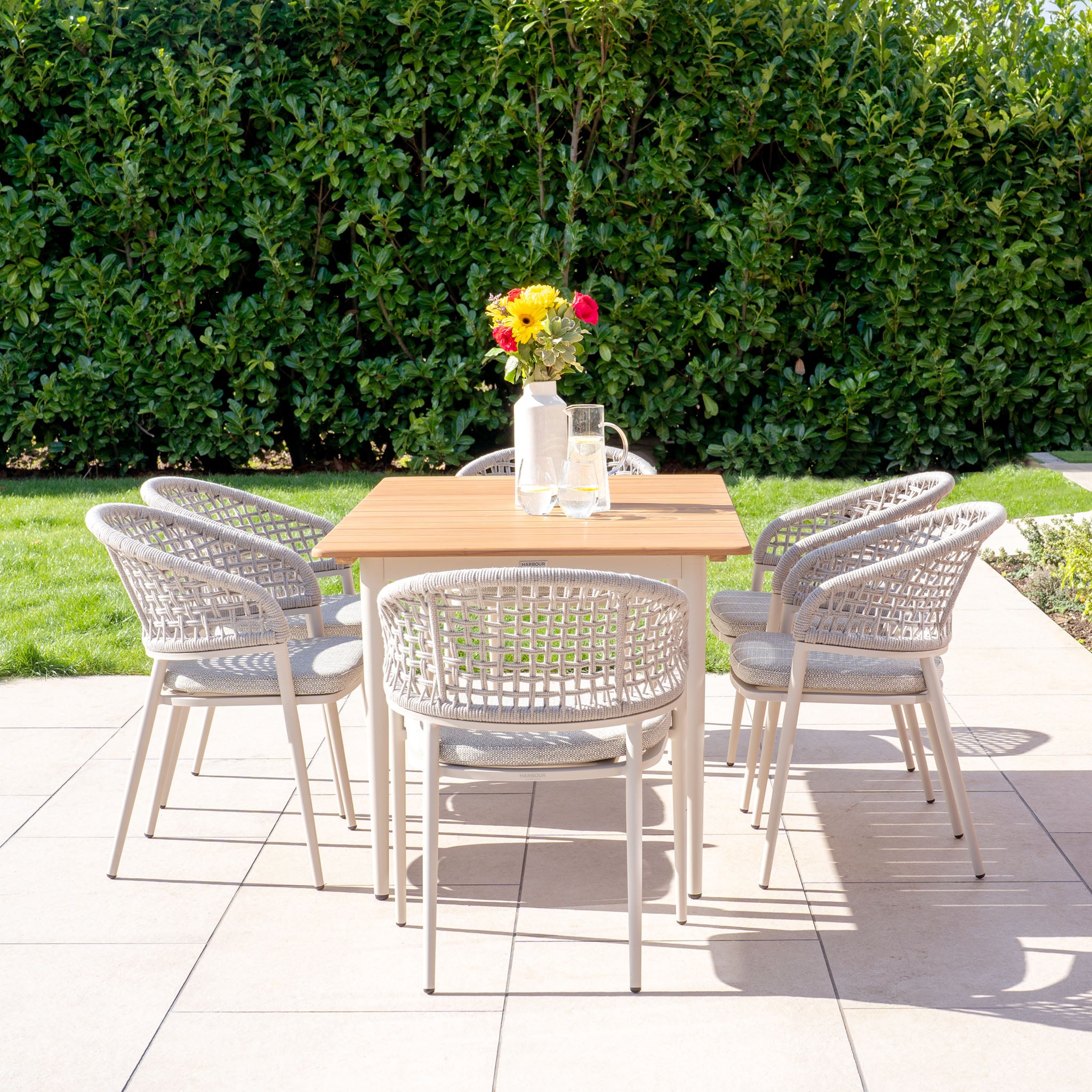 The Kalama 6 Seat Rectangular Dining Set with Teak Table in Latte features a teak outdoor dining table paired with six elegant chairs on a patio, enhanced by a flower vase and glass pitcher. This stylish ensemble is perfectly set against the backdrop of a lush green hedge.