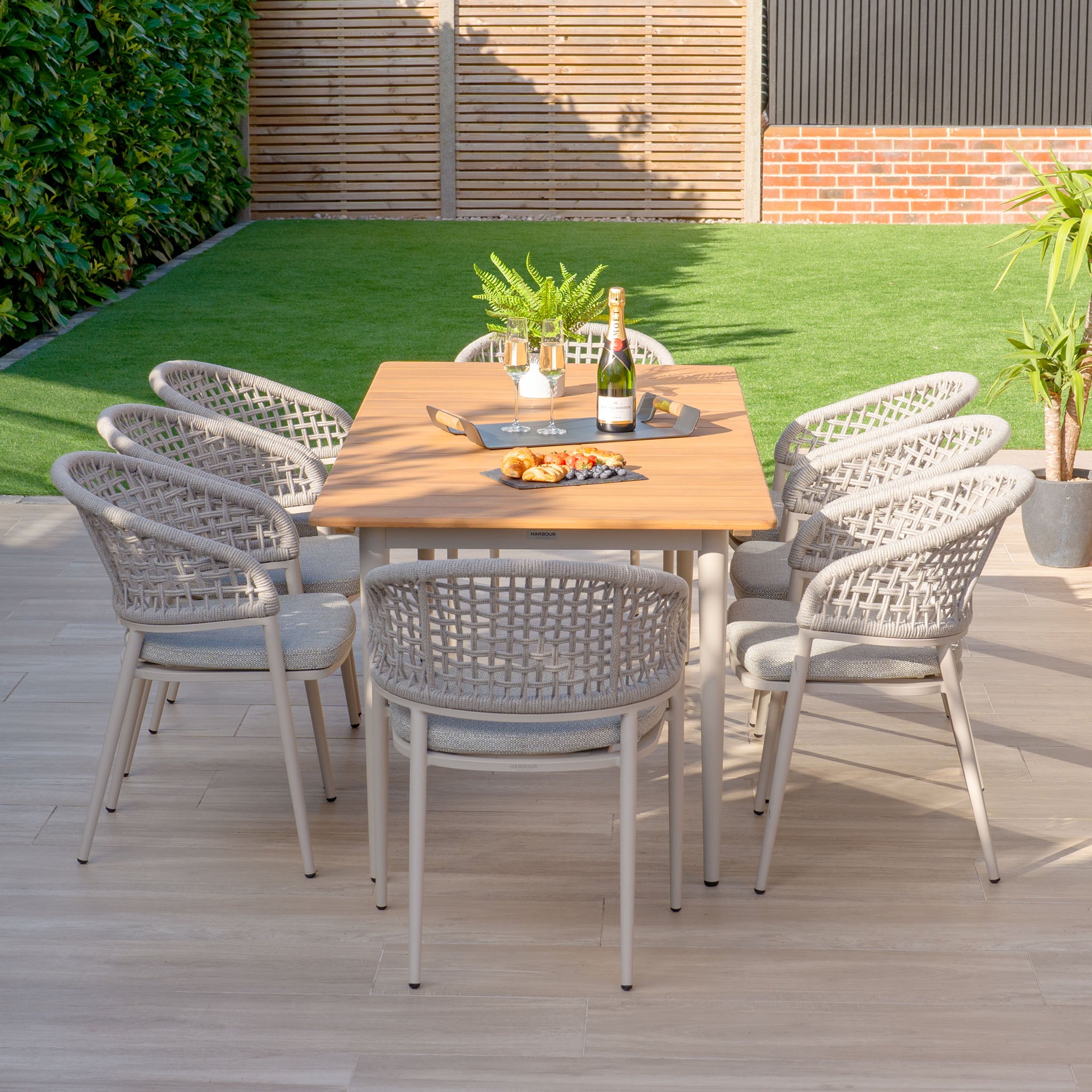 The Kalama 8 Seat Rectangular Dining Set with a Teak Table in Latte includes eight wicker chairs, all beautifully crafted with weatherproof aluminum. Ideal for outdoor get-togethers, this set is perfectly arranged on the patio with snacks and a bottle of champagne.