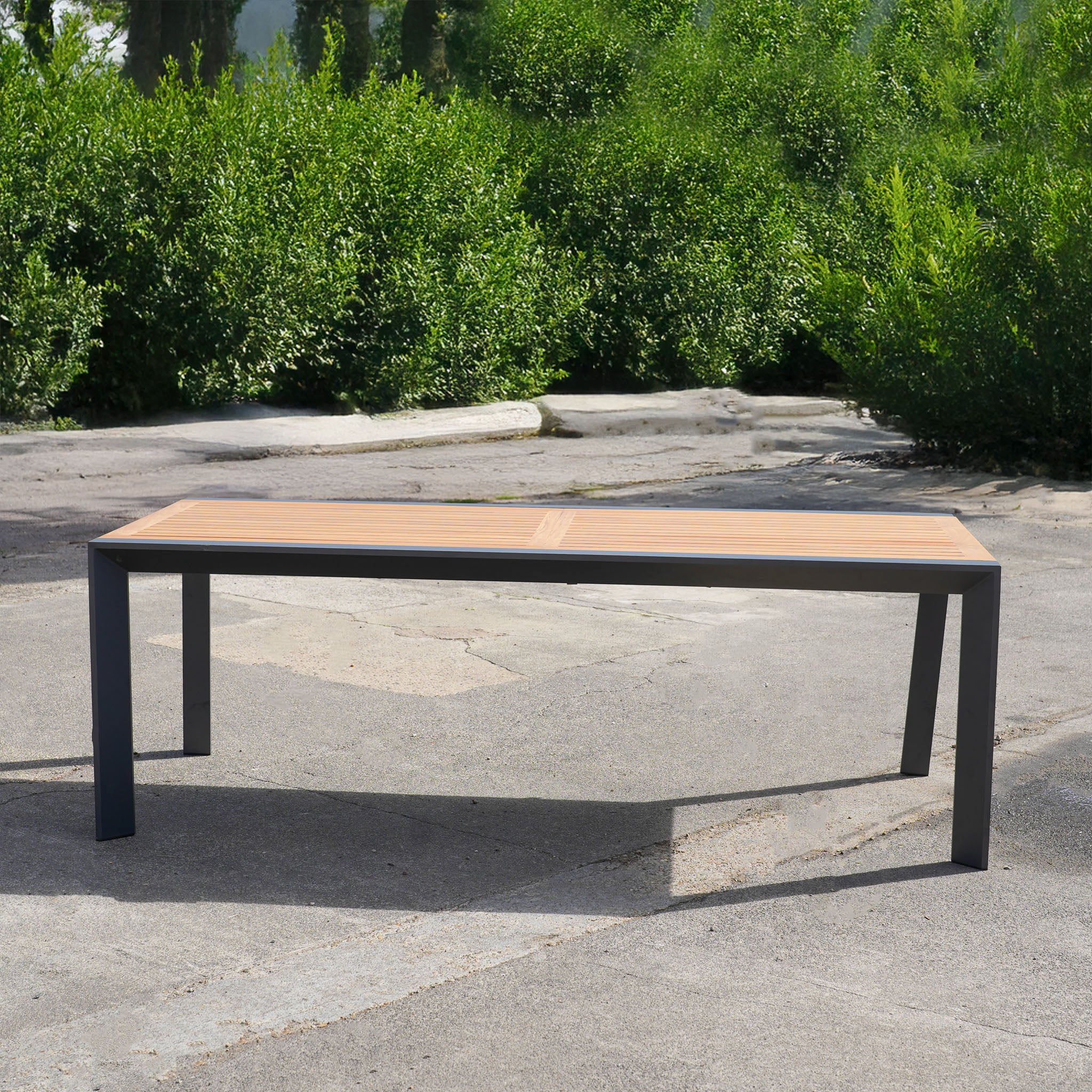 The Pier Extending Dining Table with Teak Top (Ex Display) features a modern design with sleek aluminium legs, shown outdoors on concrete and framed by lush green bushes.