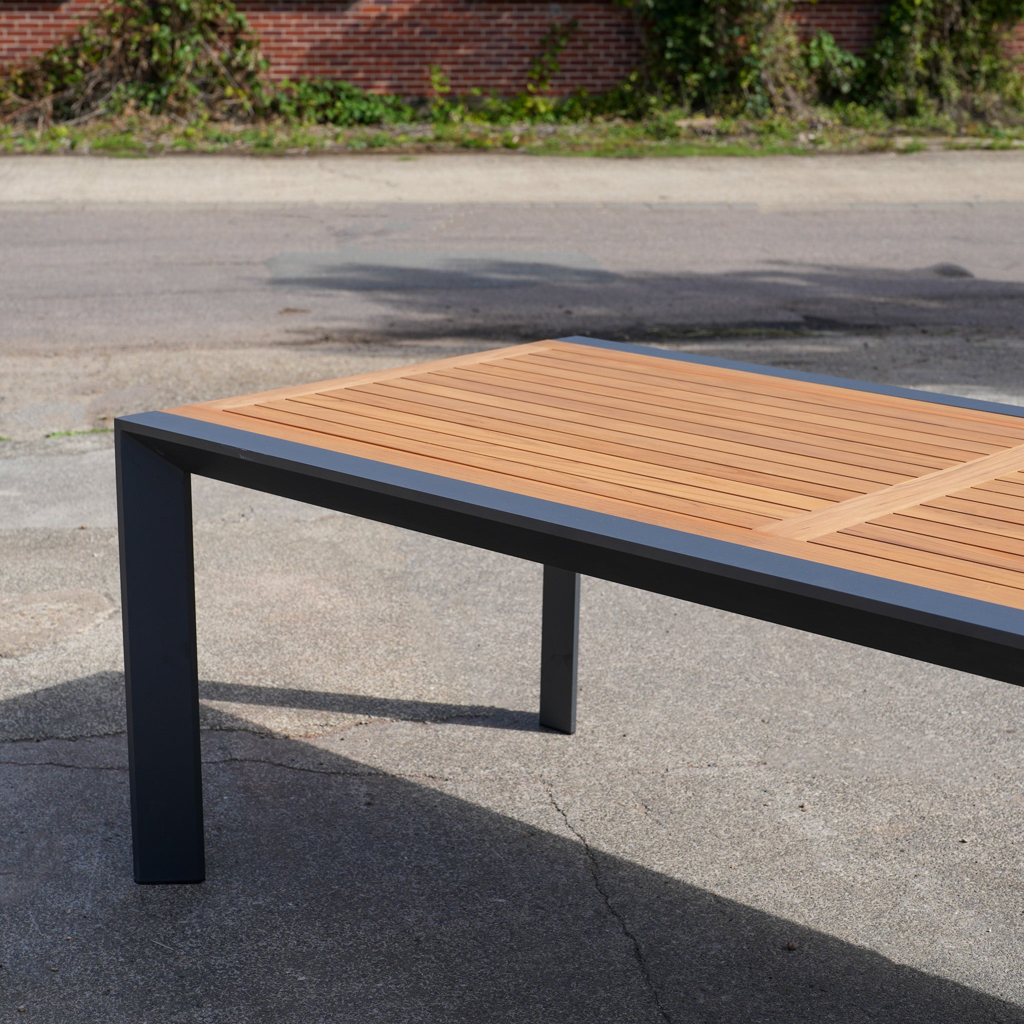 The Pier Extending Dining Table with Teak Top (Ex Display) features a teak wooden top and black aluminium legs, making it an ideal choice for outdoor dining on concrete surfaces.