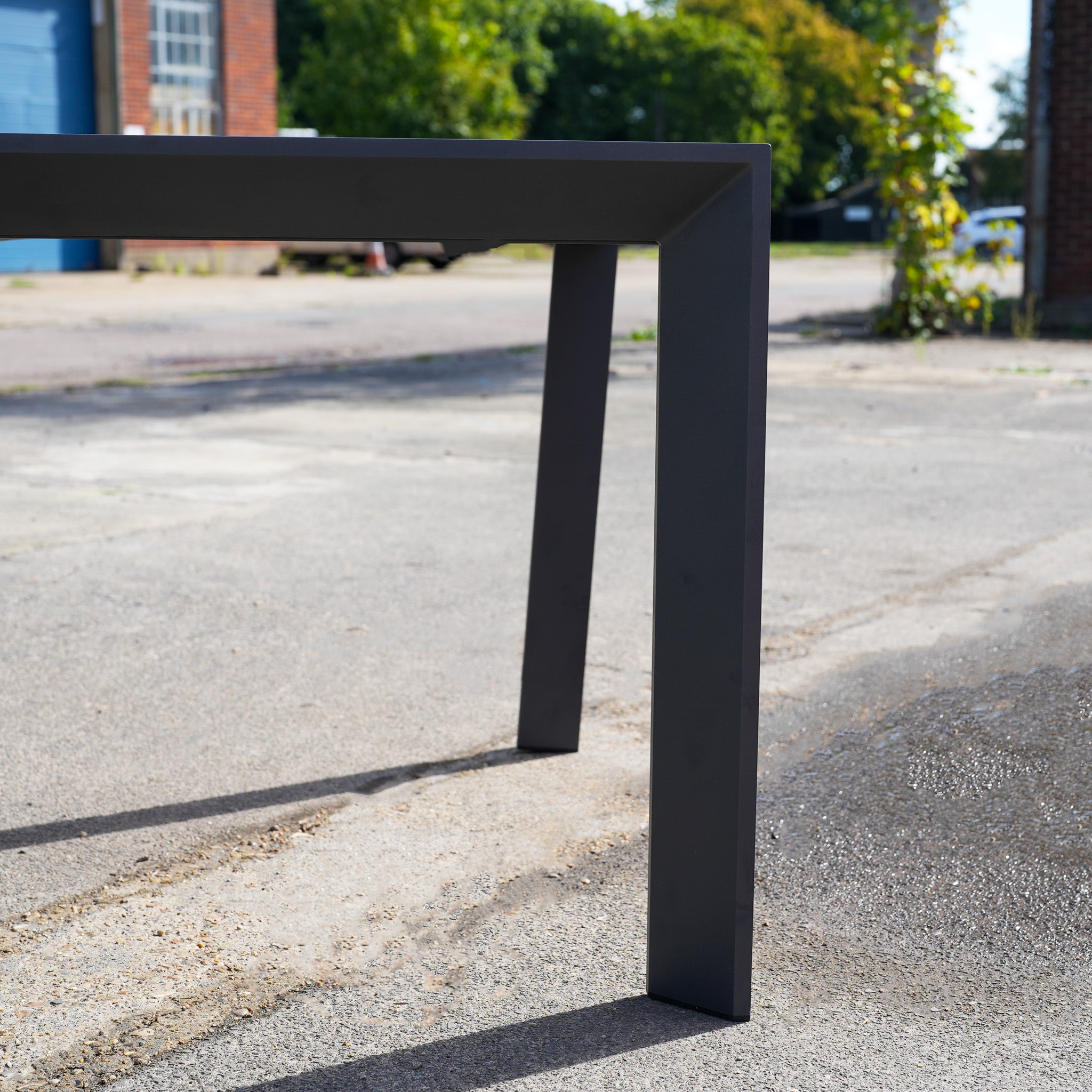 A close-up of the Pier Extending Dining Table with Teak Top (Ex Display), featuring black aluminium legs, on a sunlit outdoor pavement.