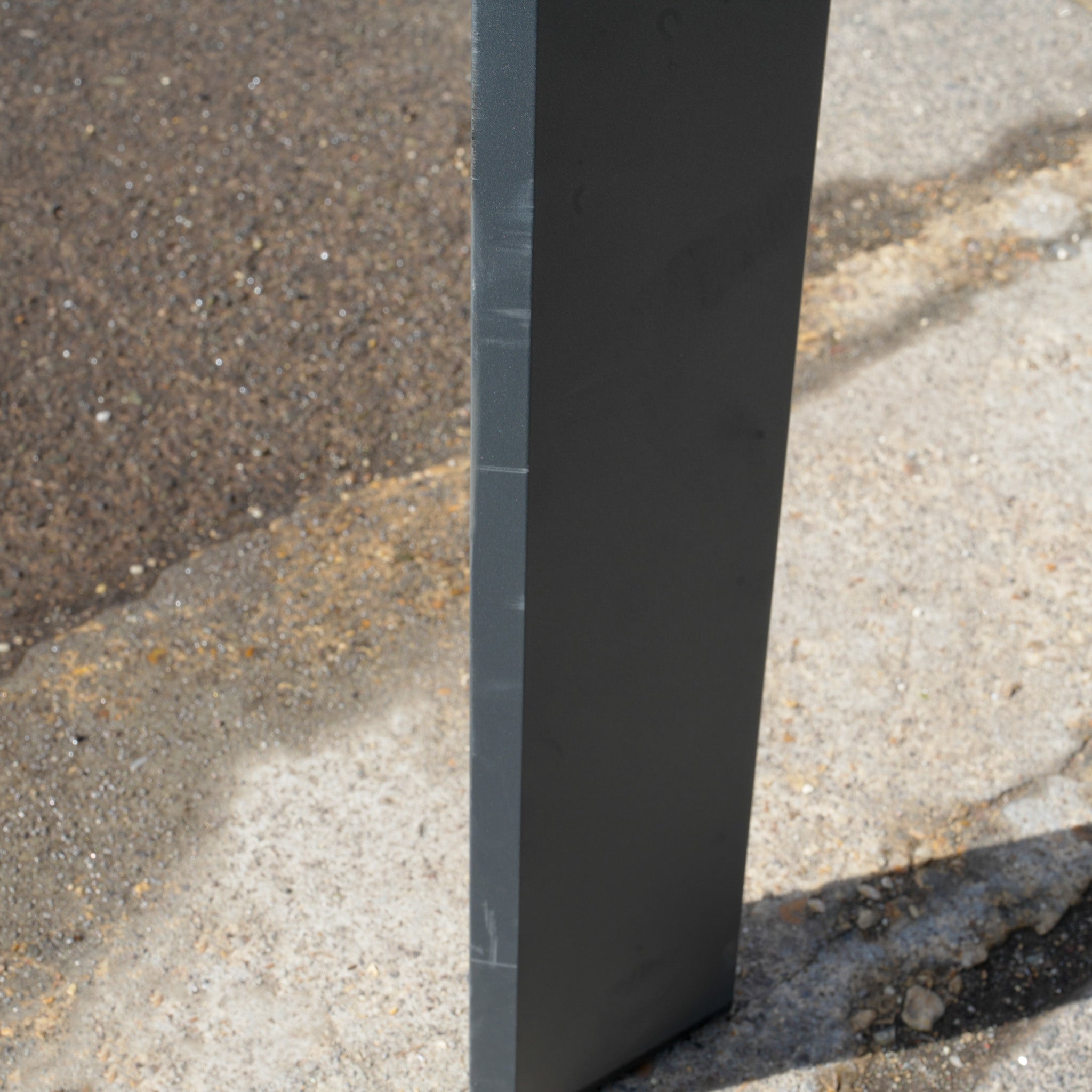 A rectangular gray metal post stands on a concrete sidewalk by an asphalt road, resembling the sturdy construction of the Pier Extending Dining Table with Teak Top (Ex Display).
