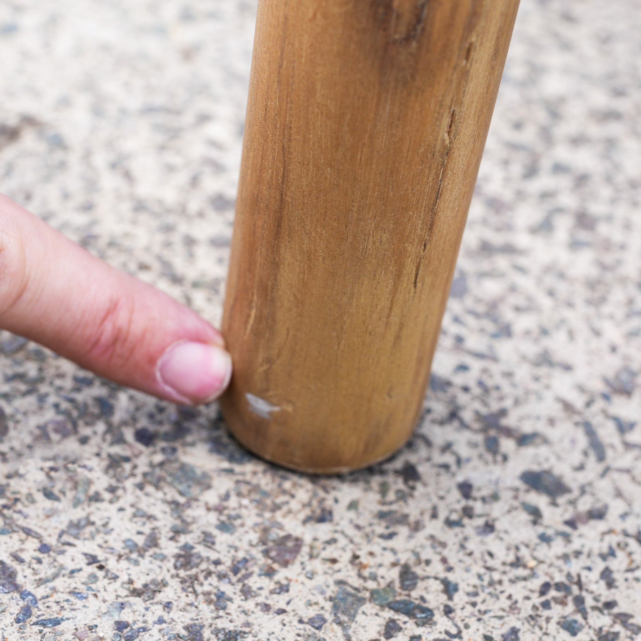 A finger points to a small chip on the bottom of a chair leg from the Paia 8 Seat Round Dining Set (Ex Display), which is resting on a speckled floor.
