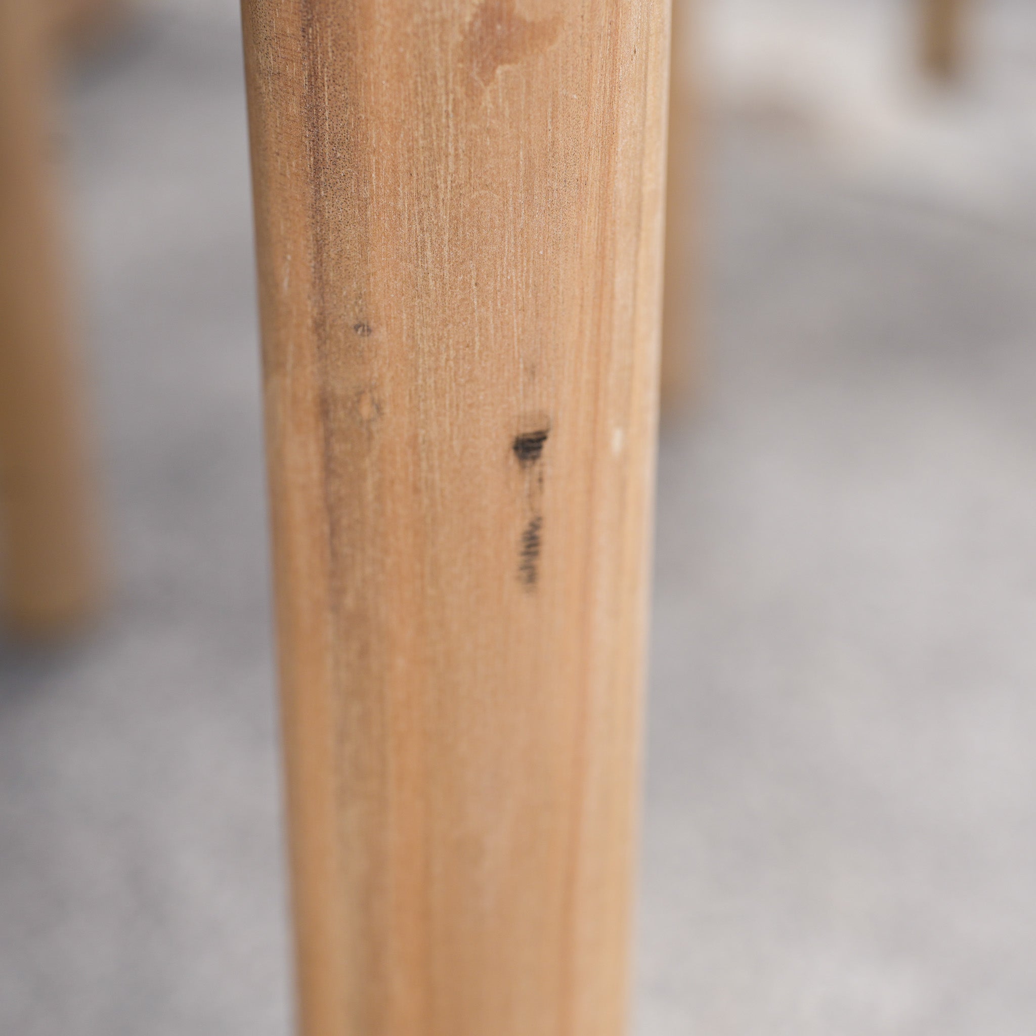 A close-up of a Paia 8 Seat Round Dining Set (Ex Display) table leg reveals acacia wood craftsmanship with a small dark mark, set against a blurred gray background.