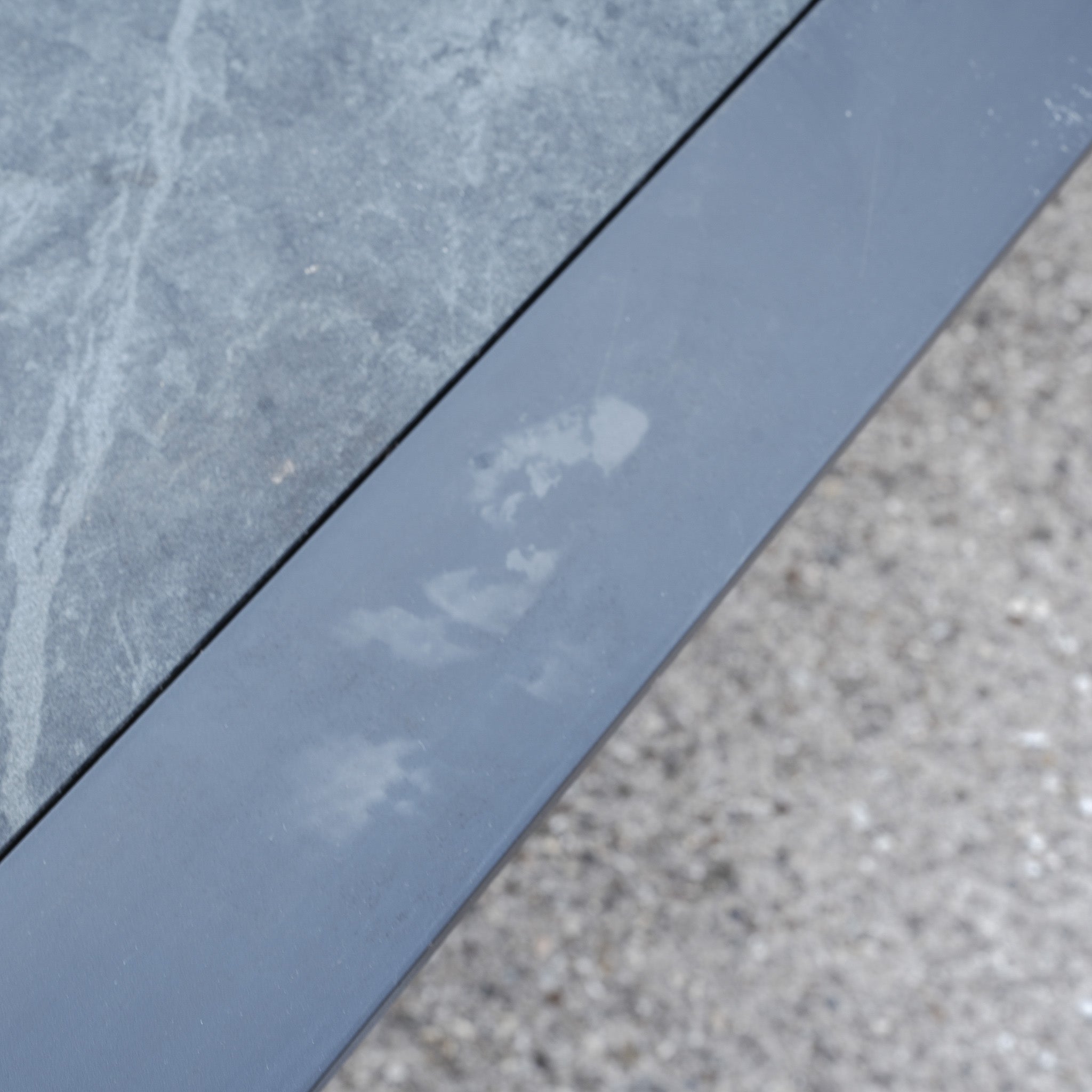 A faint white handprint on gray metal beside a glass panel and rough concrete ground captures the industrial style and sleek finish of the Pier Extending Dining Table with Ceramic Top (Ex Display).