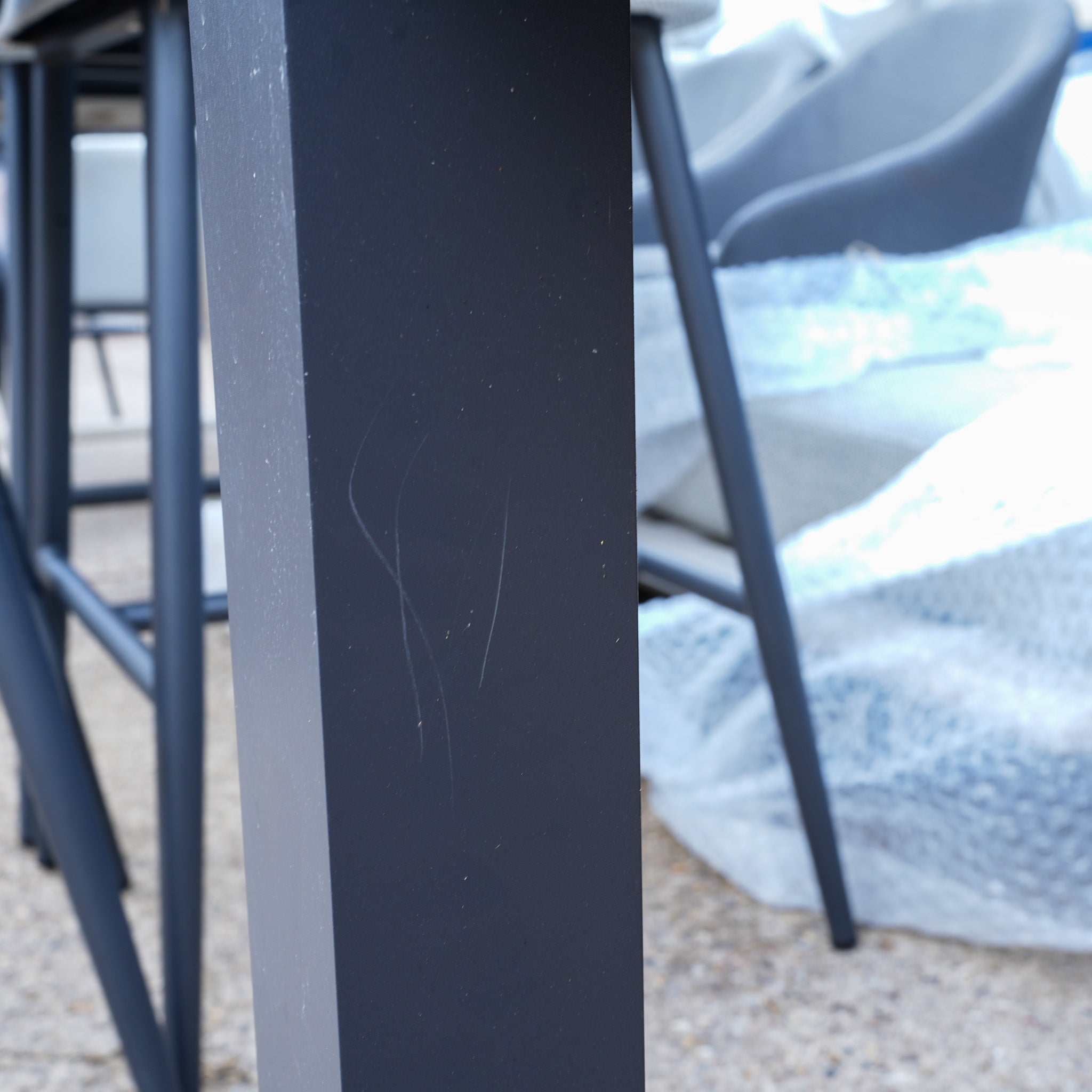 Close-up of a black metal table leg with scratches, outdoor chairs, and bubble wrap in the background—part of the Pier Extending Dining Table with Ceramic Top (Ex Display).