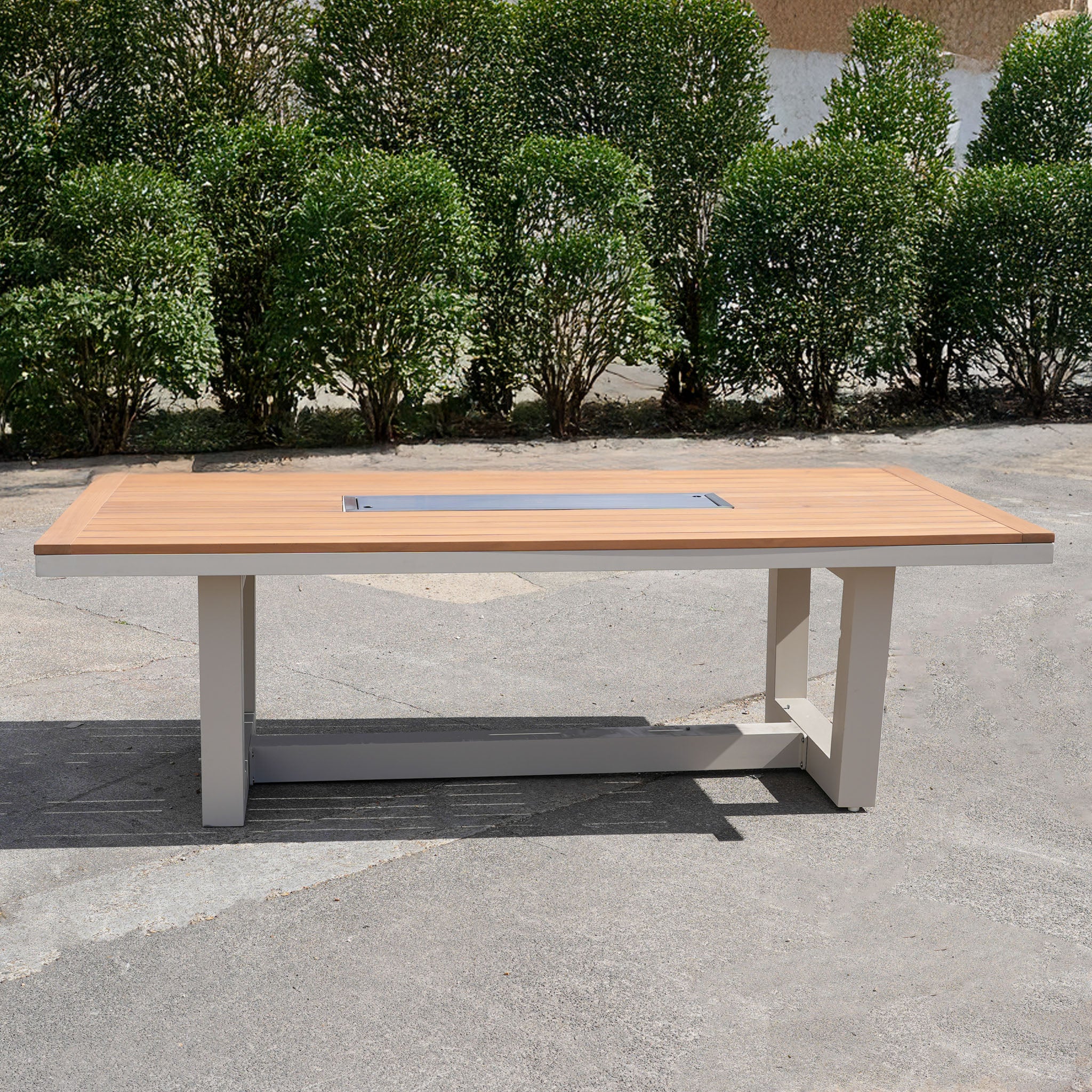 The Cloverly 8 Seat Rectangular Teak Firepit Dining Set in Latte (Ex Display) is placed outdoors in front of green bushes.