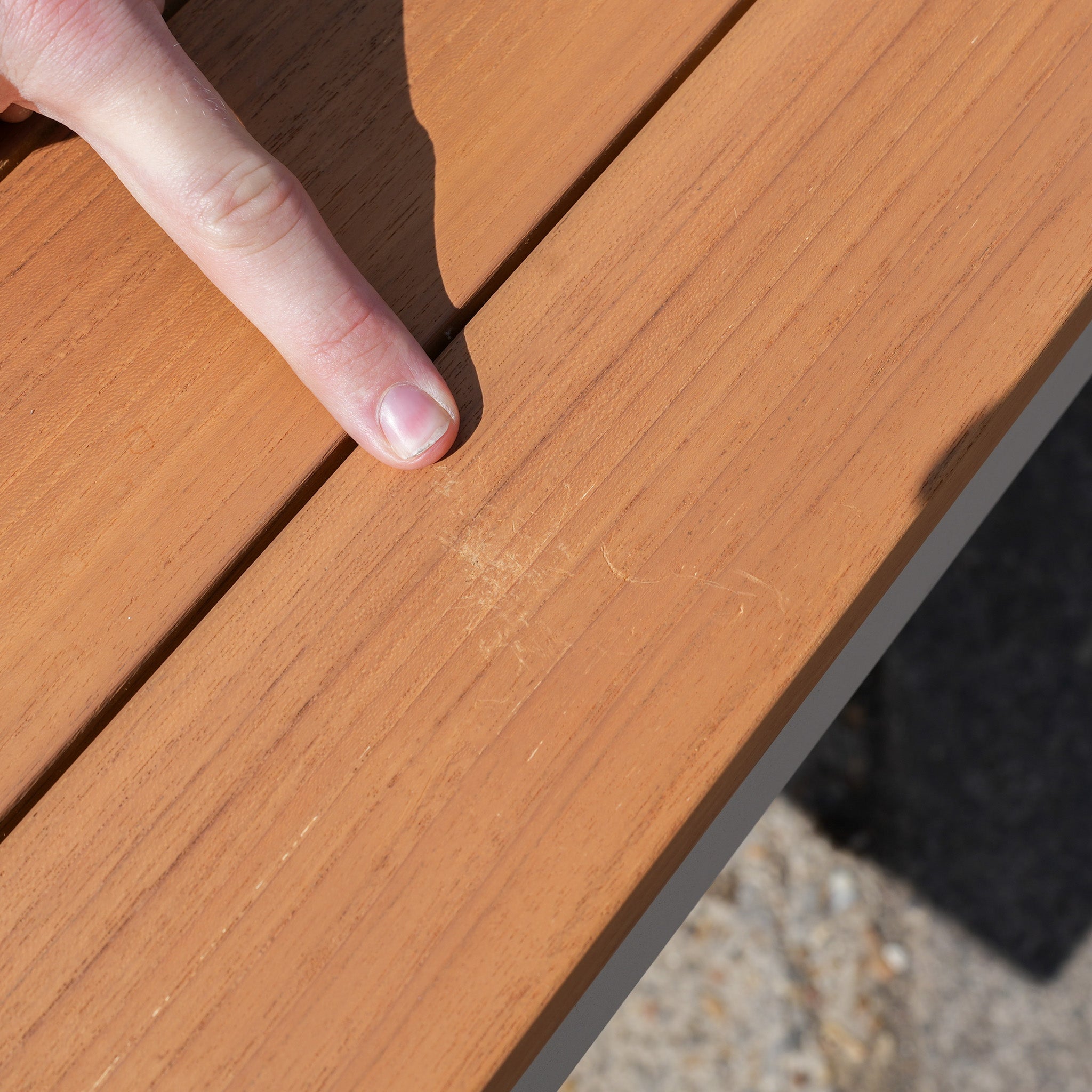 A finger points to a small scratch on a wooden surface in bright sunlight, next to the Cloverly 8 Seat Rectangular Teak Firepit Dining Set in Latte (Ex Display).