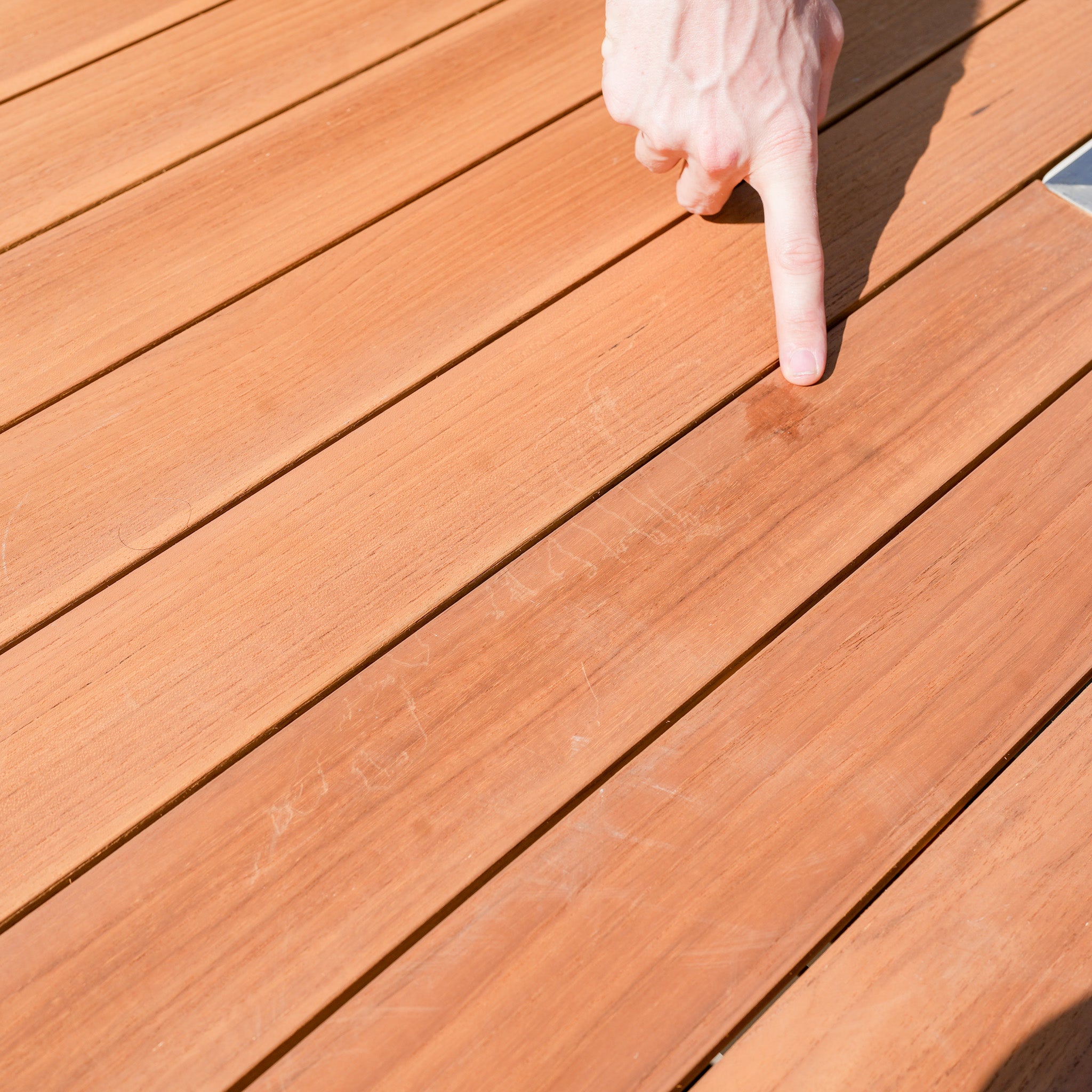 A hand points to a scratch on a wooden deck in bright sunlight, next to the Cloverly 8 Seat Rectangular Teak Firepit Dining Set in Latte (Ex Display).