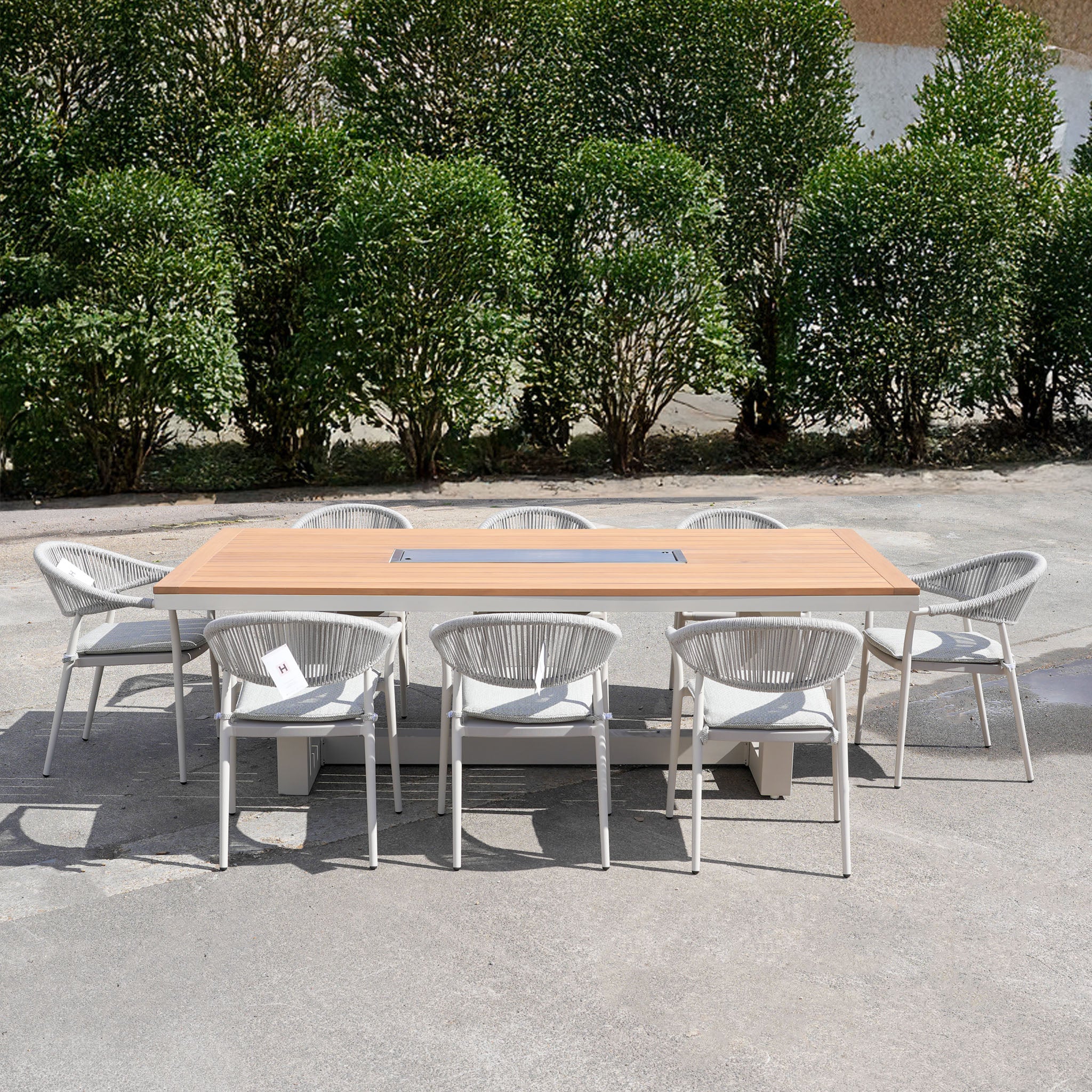 The Cloverly 8 Seat Rectangular Teak Firepit Dining Set in Latte (Ex Display) is shown outdoors, featuring eight chairs around a teak table on a patio with green hedges in the background.