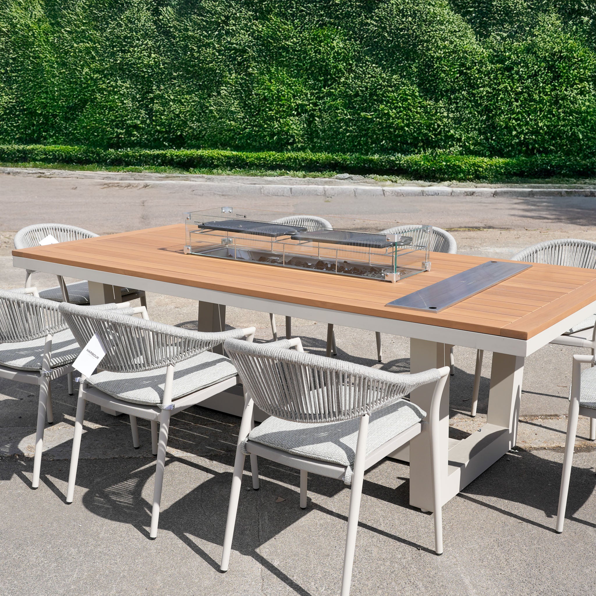 The Cloverly 8 Seat Rectangular Teak Firepit Dining Set in Latte (Ex Display) features a wooden table and eight rope chairs—ideal for stylish outdoor gatherings next to a green hedge.