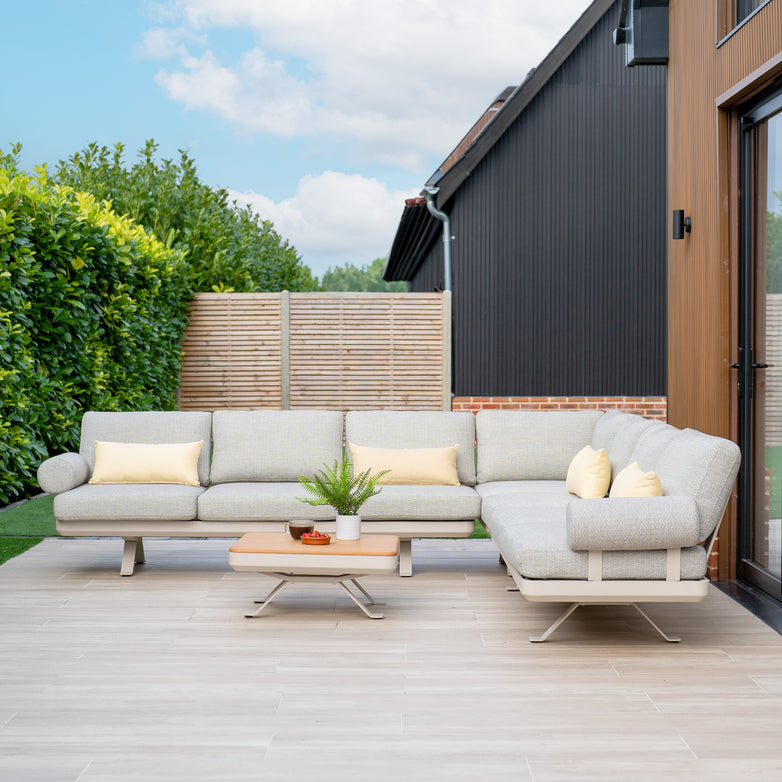 Outdoor patio featuring the Vienna Corner Group Set in Latte, accented with yellow pillows, a coffee table, and lush green plants in the background.