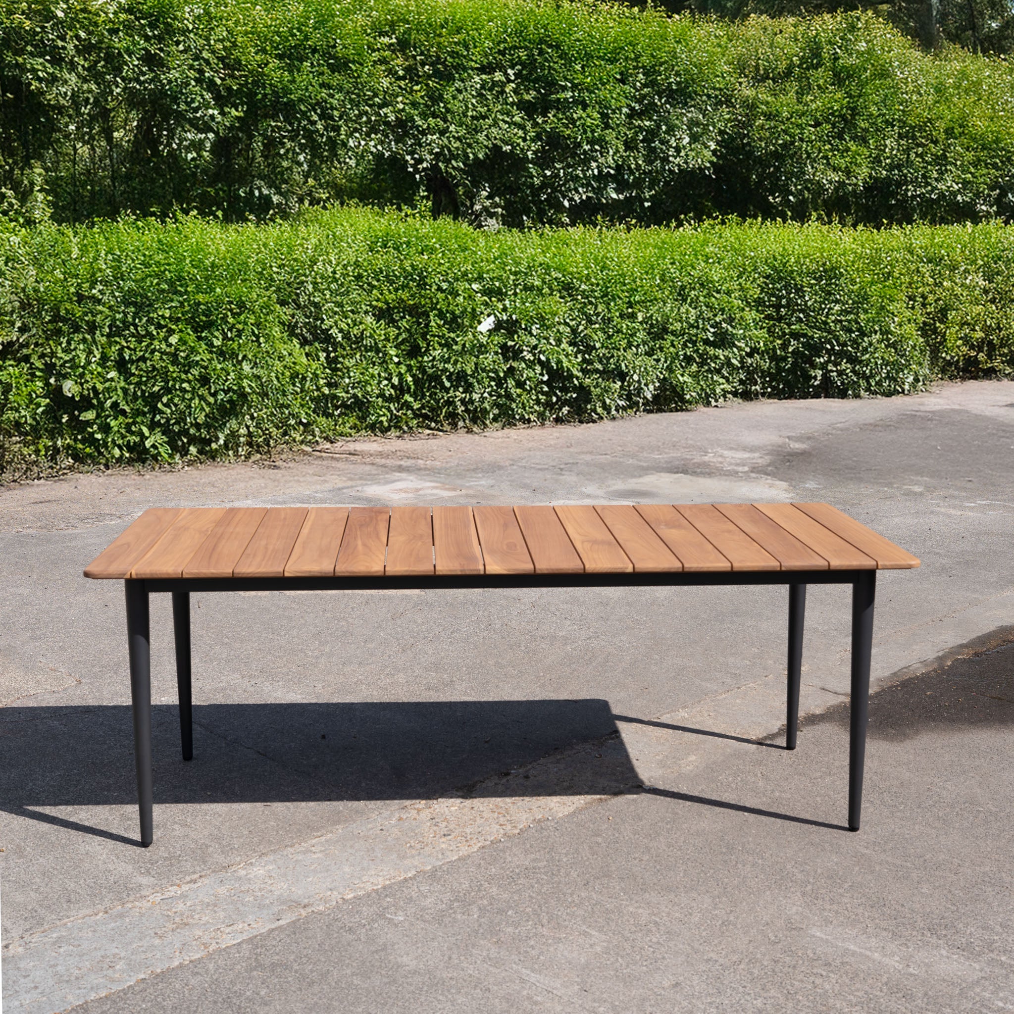 The Bay 8 Seat Dining Table in Charcoal (Ex Display) features a golden teak top and black metal legs, set on pavement before green hedges—ideal as a stylish teak outdoor dining table.