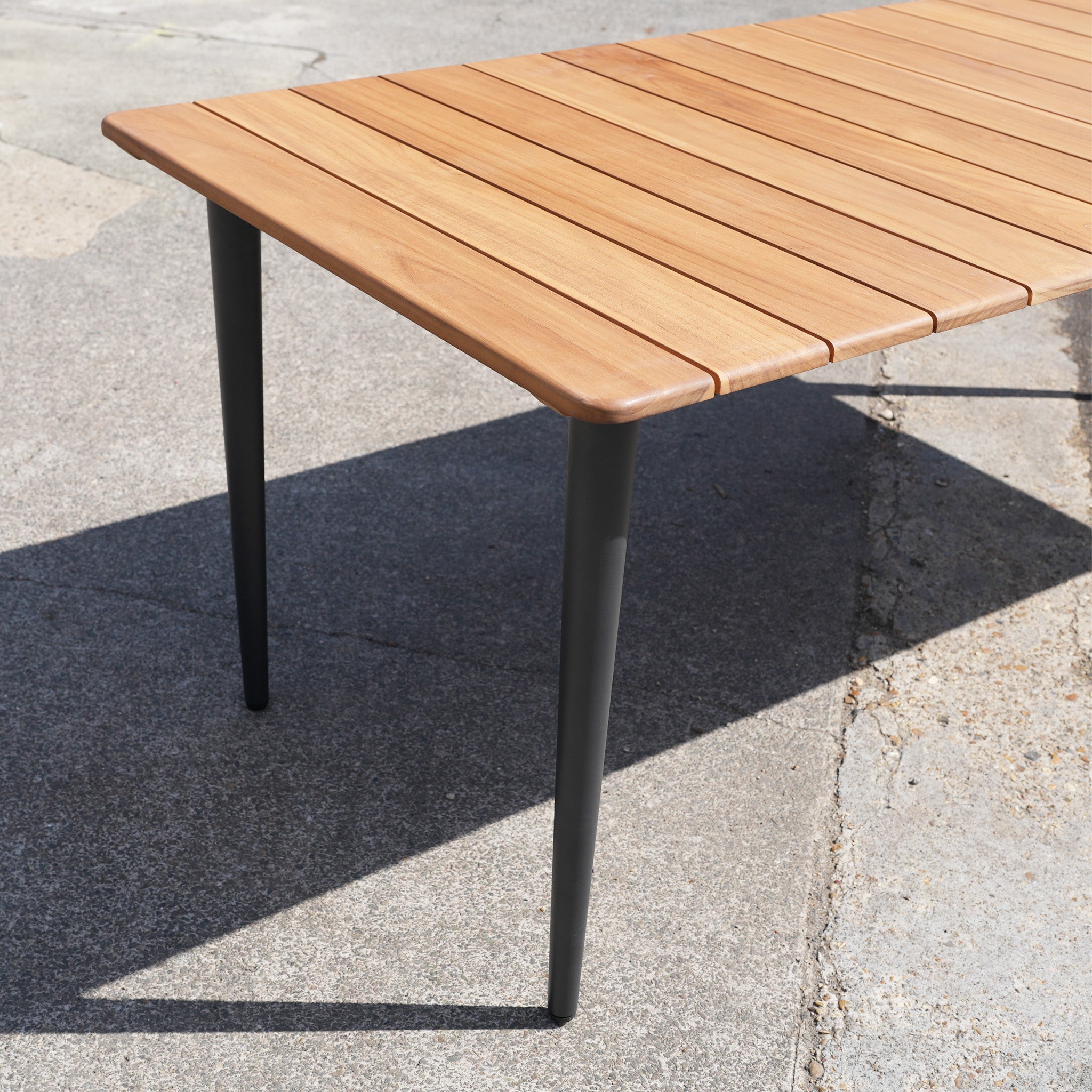 The Bay 8 Seat Dining Table in Charcoal (Ex Display) features a golden teak top with black metal legs, ideal for outdoor dining. Place it on concrete in sunlight and follow the wooden care guide to keep it looking beautiful.