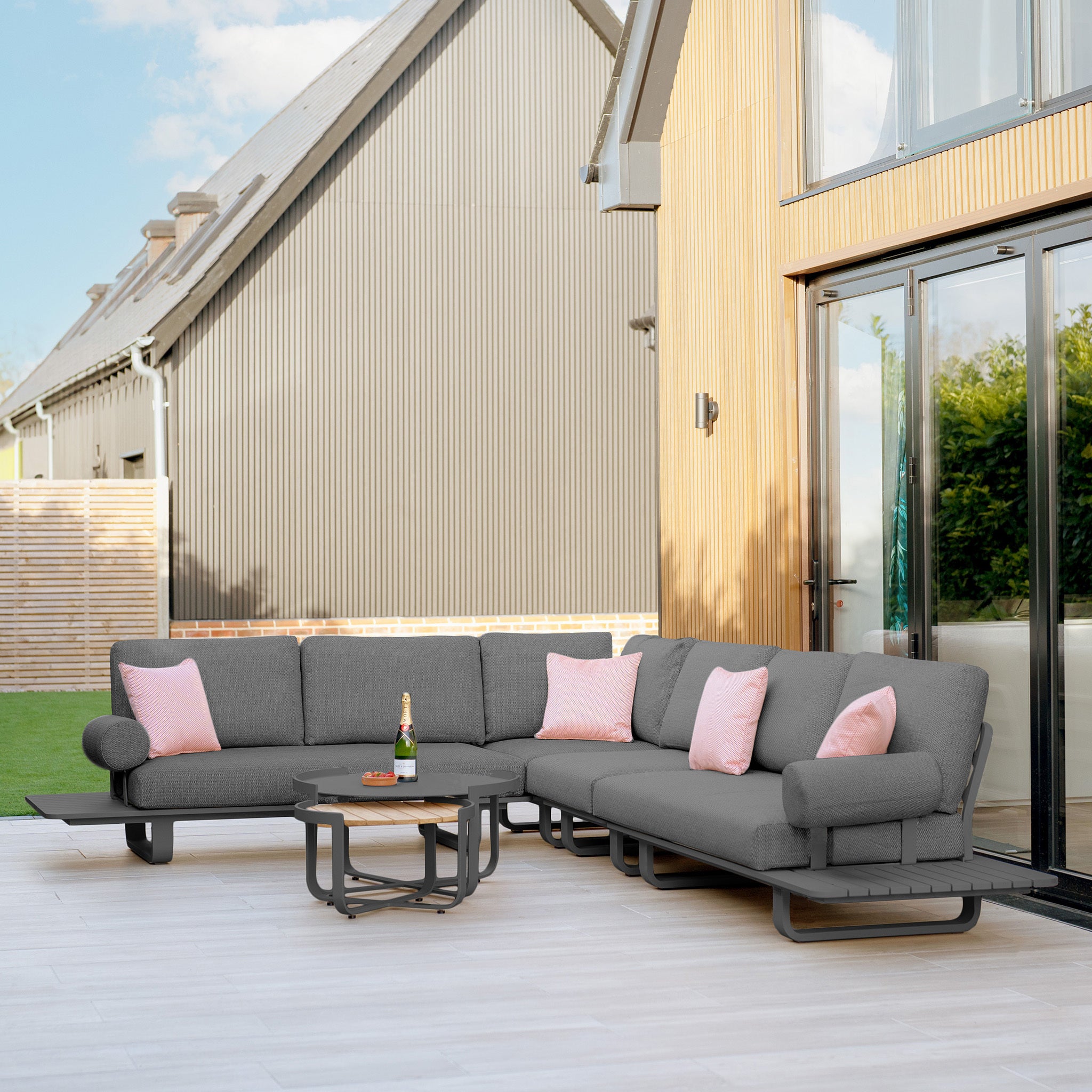 Outdoor patio with a Bay Large Corner Group Set with an aluminum frame in charcoal, showcasing gray sectional seating and UV-resistant Olefin pink pillows. A teak table top supports a small table and a bottle, soaking up the sunlight next to the house.