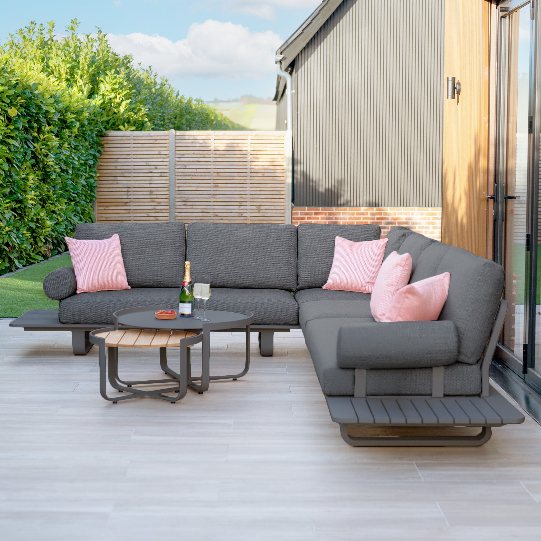 The Bay Large Corner Group Set with an aluminium frame in charcoal pairs with a round teak coffee table, drinks, and greenery to create a modern patio setting.