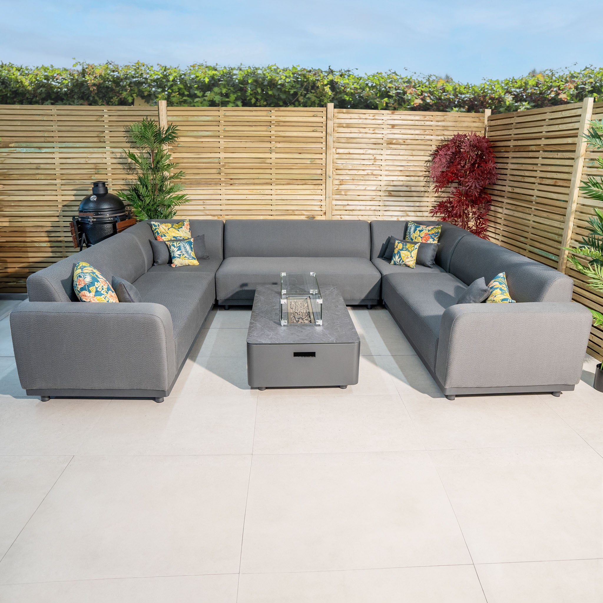 The Luna U-Shape Outdoor Fabric Sofa Set with Firepit Coffee Table in Grey, adorned with colorful throw pillows, is the centerpiece of the patio.