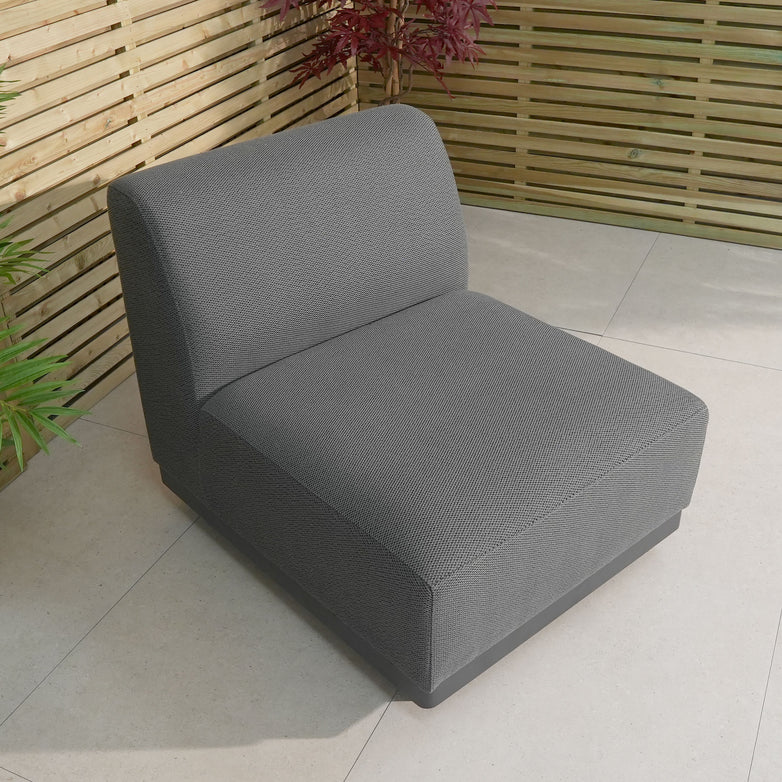 The Luna Outdoor Fabric Middle Extension Piece in Grey is an elegant patio lounge option with quick-dry foam cushions. It complements a tiled patio with wooden slat fencing and vibrant potted plants, ideal for outdoor fabric enthusiasts.