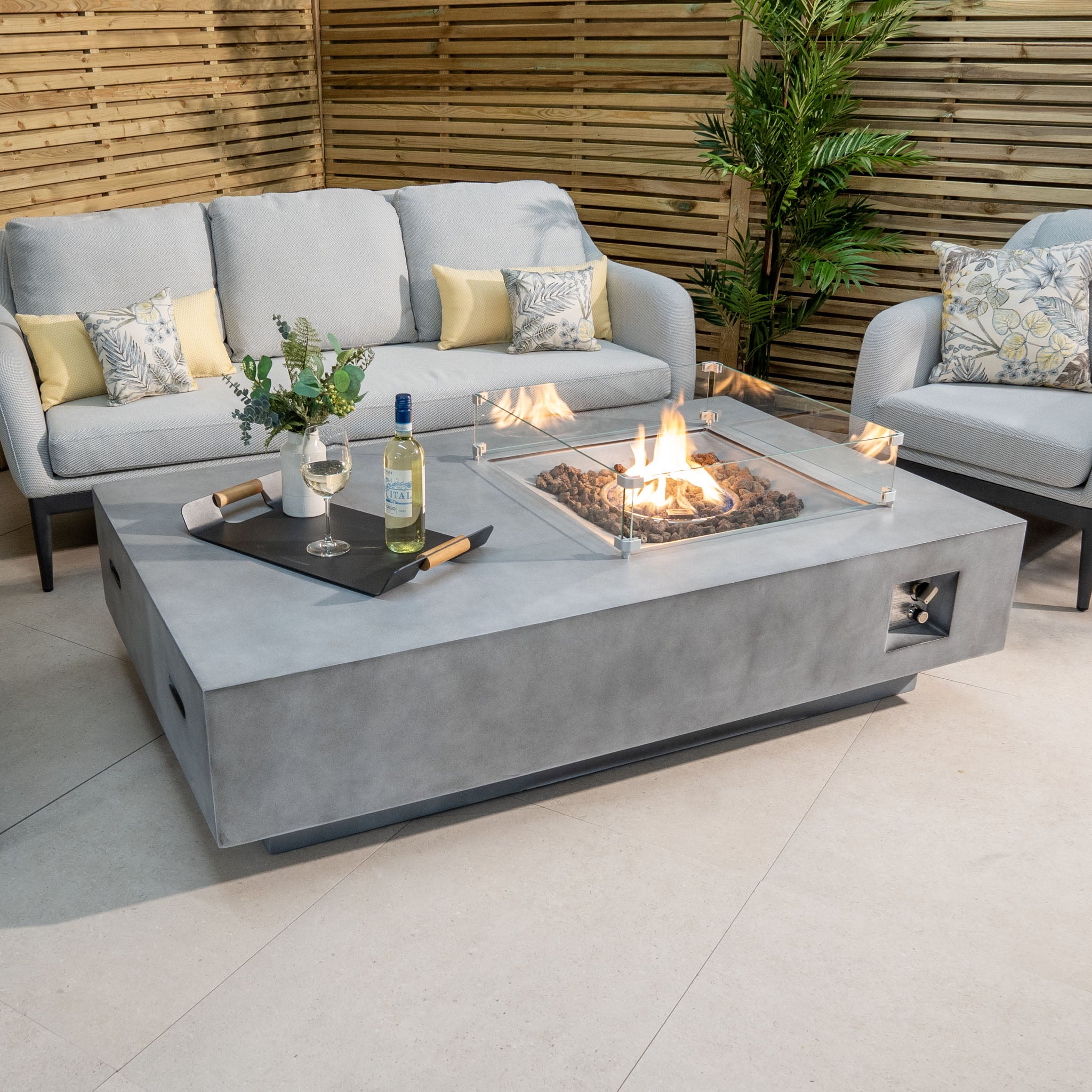 Outdoor seating area with a sofa, armchairs, and an Ember Rectangular Firepit Coffee Table in Stone Grey boasting a 14kW heat output. Glasses of wine rest on a tray beside the modern fire pit made from 304 stainless steel.
