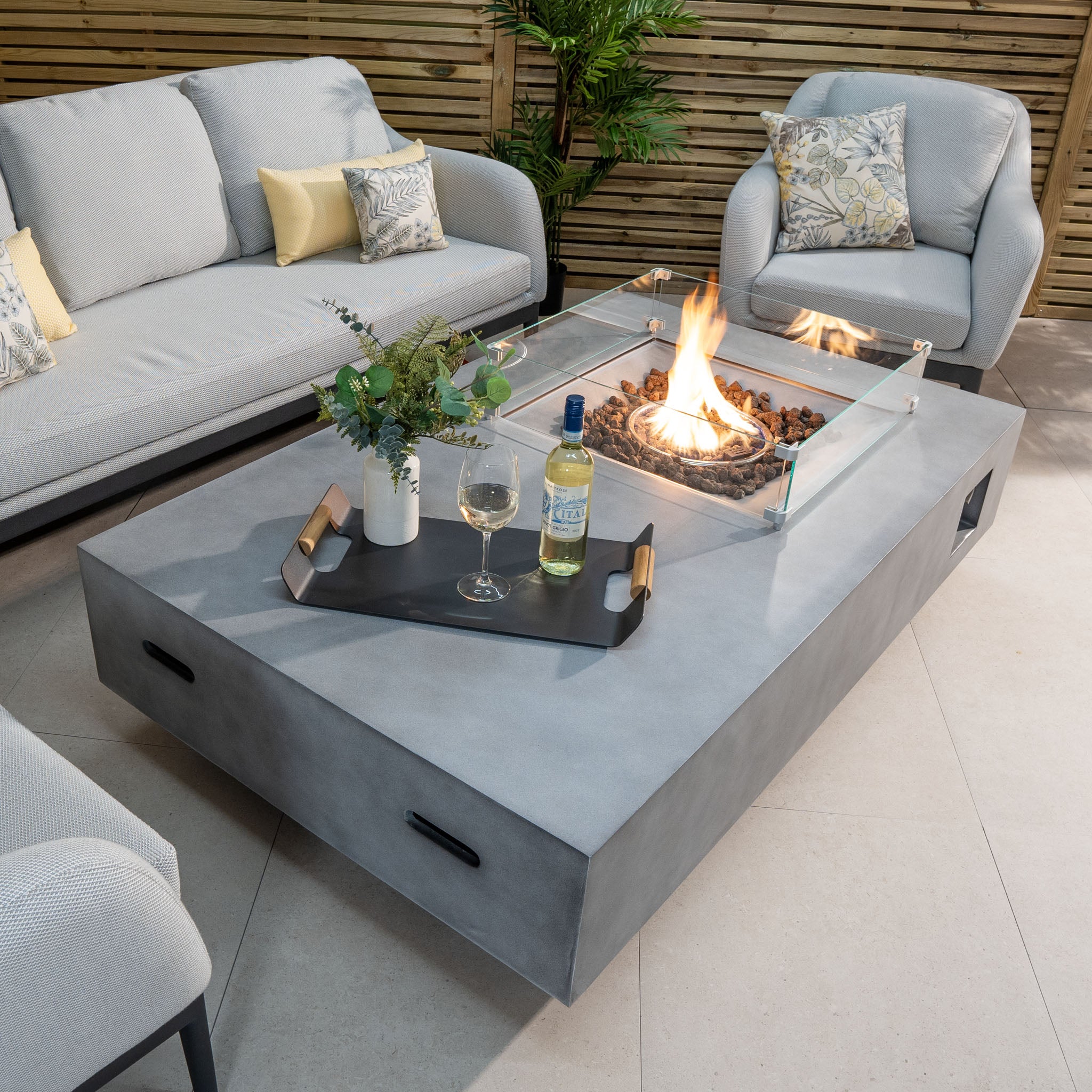 Modern outdoor seating area with a 14kW heat output from the Ember Rectangular Firepit Coffee Table in Stone Grey, surrounded by cushioned sofas and chairs.