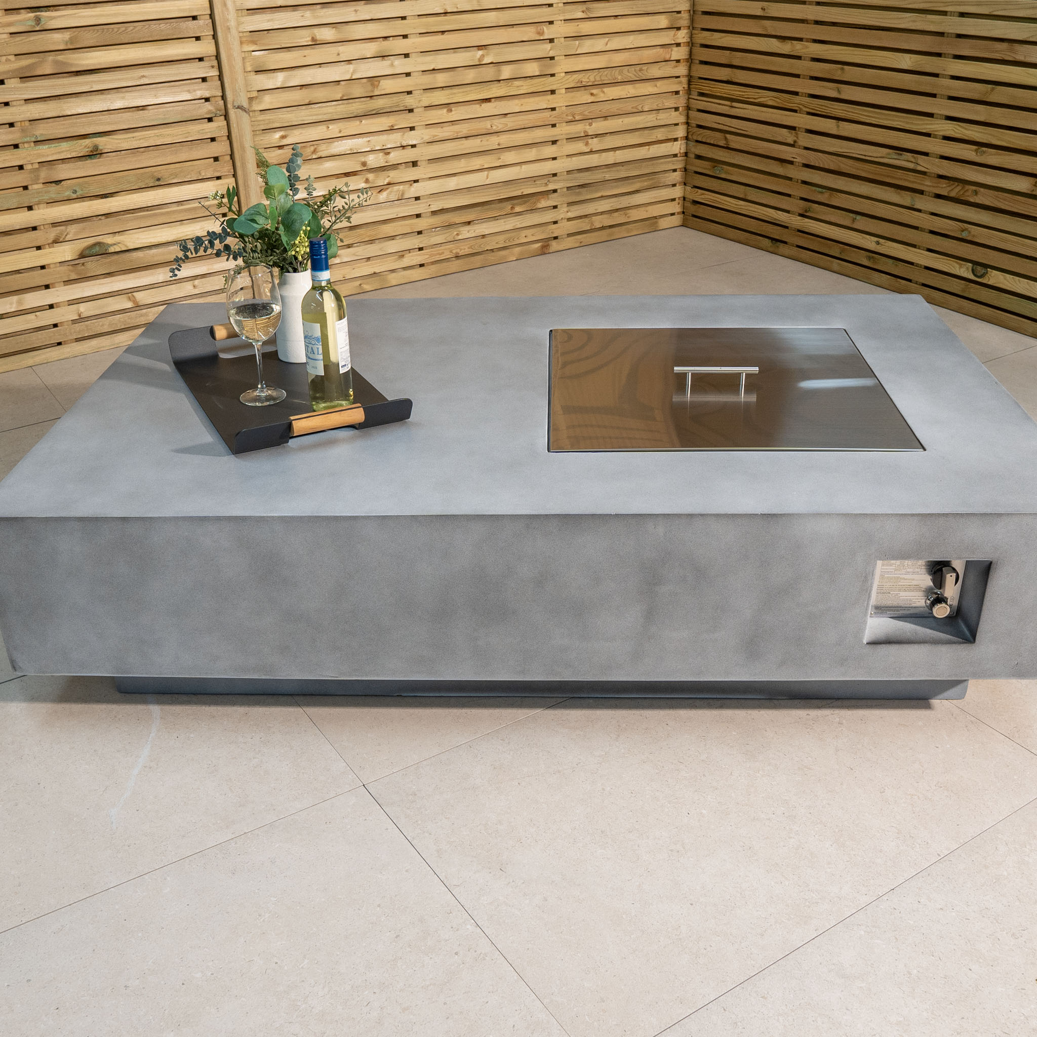 A stylish Ember Rectangular Firepit Coffee Table in Stone Grey with a 14kW heat output, featuring a concrete design and a tray holding wine bottles, glasses, and a plant arrangement on top.