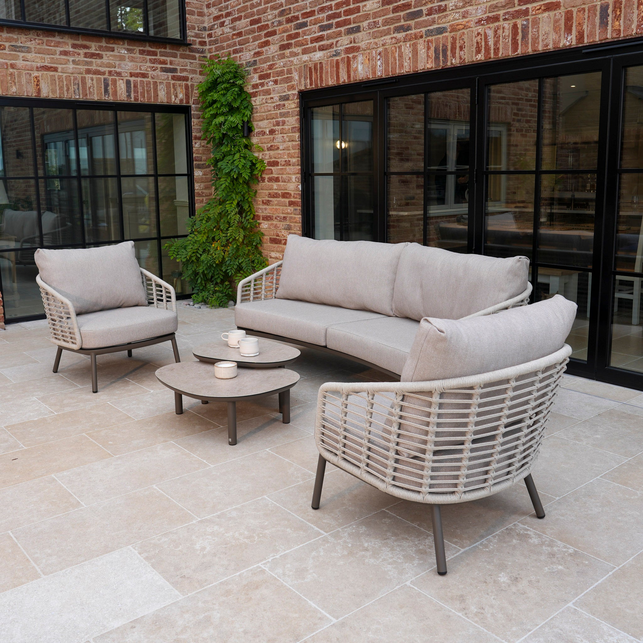 The Sutera 3 Seat Sofa Set in Putty features a sofa, two armchairs, and round tables, perfect for your patio with stone tiles near brick walls and windows.