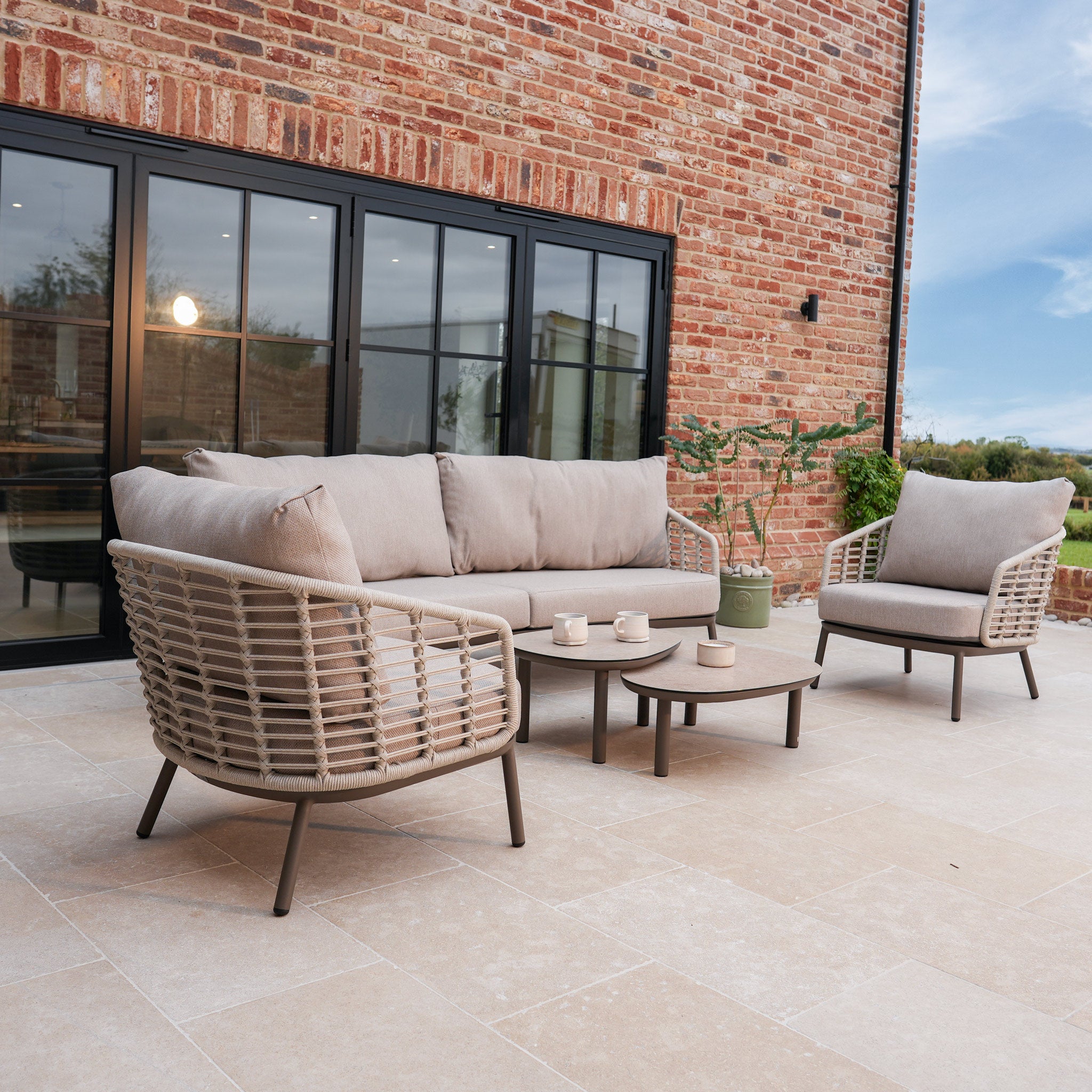 The Sutera 3 Seat Sofa Set in Putty, complete with all-weather cushions, armchairs, and a table, is arranged on a stone patio by a brick house with a potted plant.