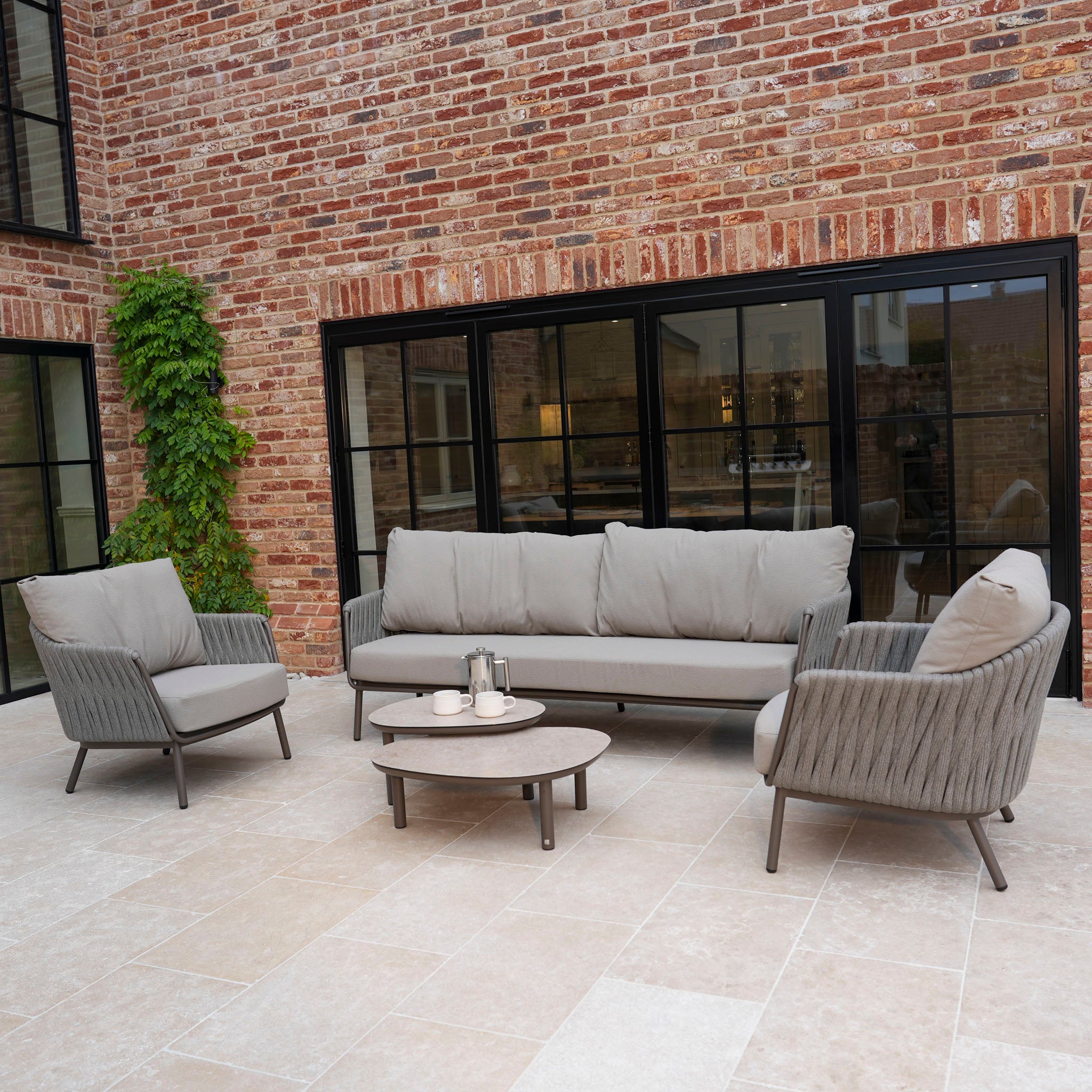 Outdoor patio featuring a Capri 3 Seat Sofa Set in Stone Grey with hand-woven rope details, two armchairs, a round coffee table, and brick walls with large windows.