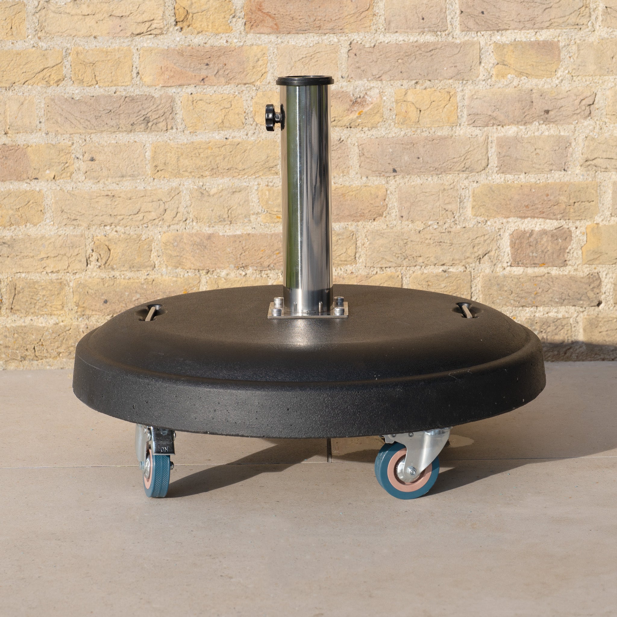 50kg Concrete Parasol Base with Handles & Wheels, featuring a metal pole, placed on pavement before a beige brick wall.