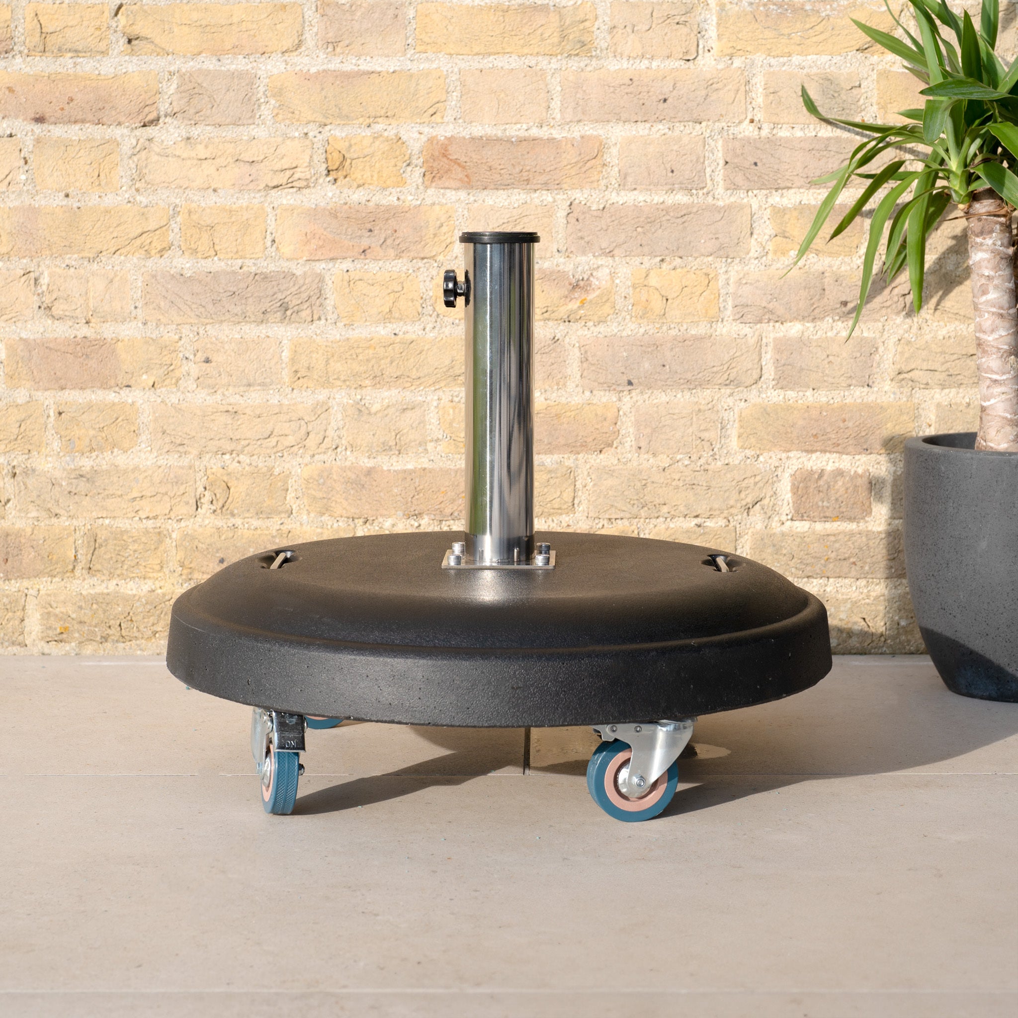 The 50kg Concrete Parasol Base with Handles & Wheels is set on the patio near a potted plant and brick wall.