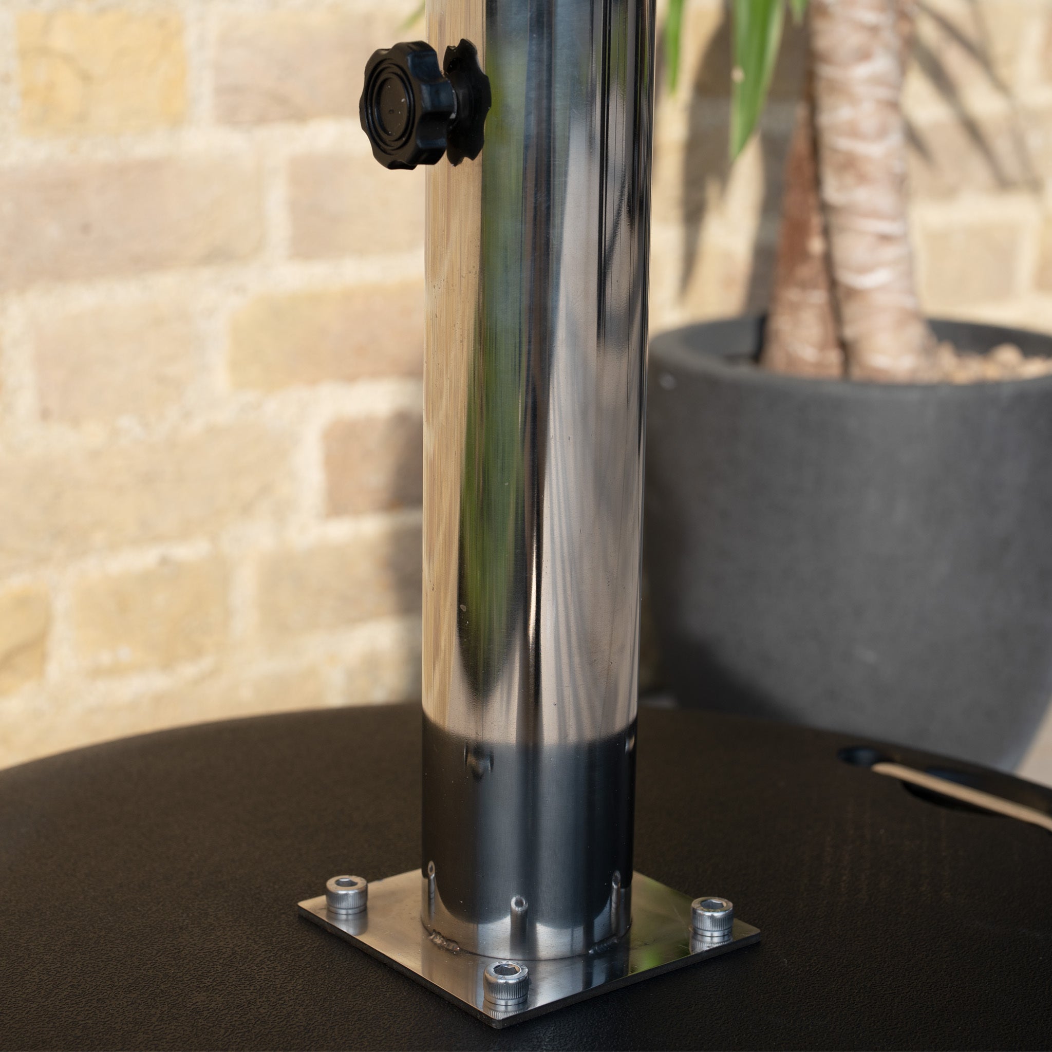 A shiny metal pole stands in a 50kg Concrete Parasol Base with Handles & Wheels, set against a background featuring a potted plant and brick wall.