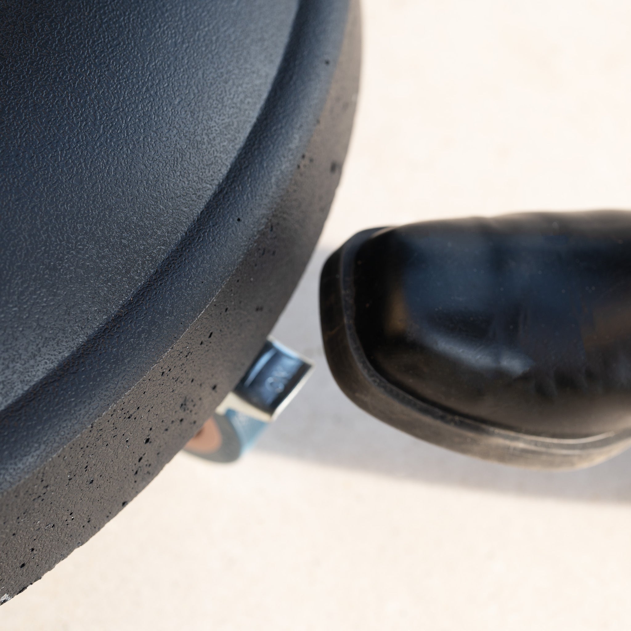 Close-up of a black chair seat with a wheel and a shoe on the base, highlighting the robust design of the 50kg Concrete Parasol Base with Handles & Wheels, set on a light surface.