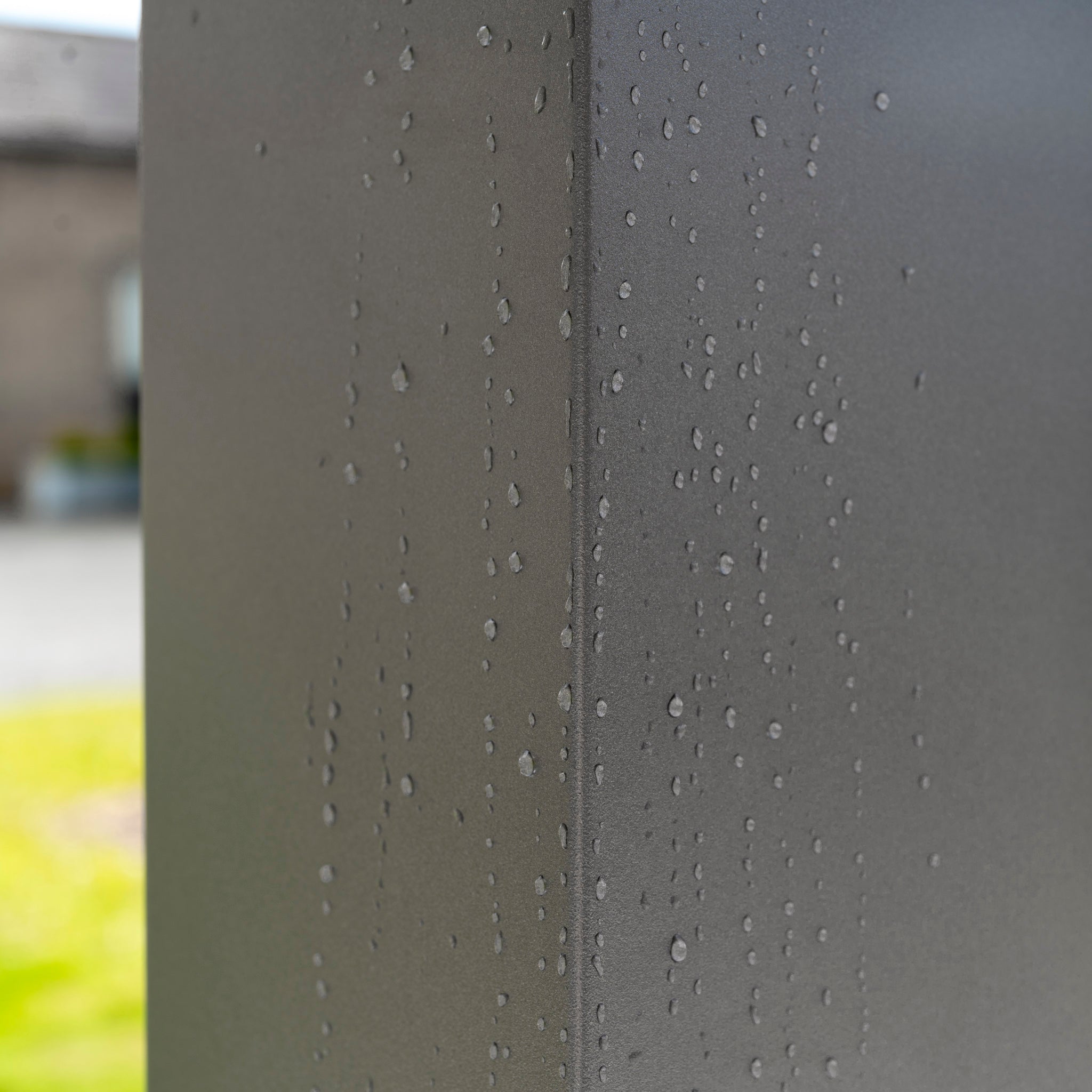 Close-up of raindrops on a sleek, gray metal surface outdoors, possibly the PergoSTET Deluxe 3m x 6m Rectangular Pergola with Motorised LED Louvres and 4 Manual Side Blinds, with blurred buildings and grass in the background.
