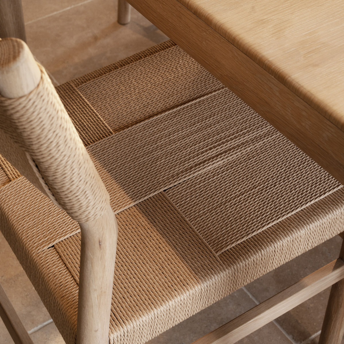 A close-up of a handwoven seat from the Marlow 8 Seat Dining Set in Light Oak, positioned beside its matching solid oak dining table on a tiled floor.